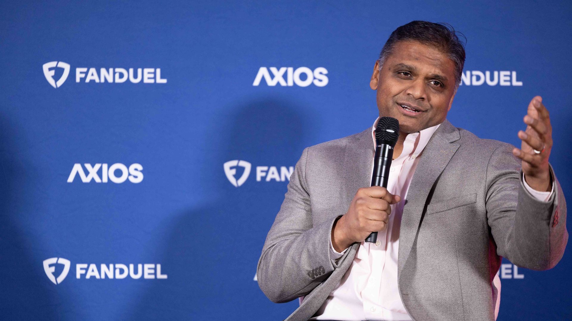 Man in gray blazer and white shirt speaking into a microphone, gesturing with his hand, seated against a blue backdrop with white "FANDUEL" and "AXIOS" logos.