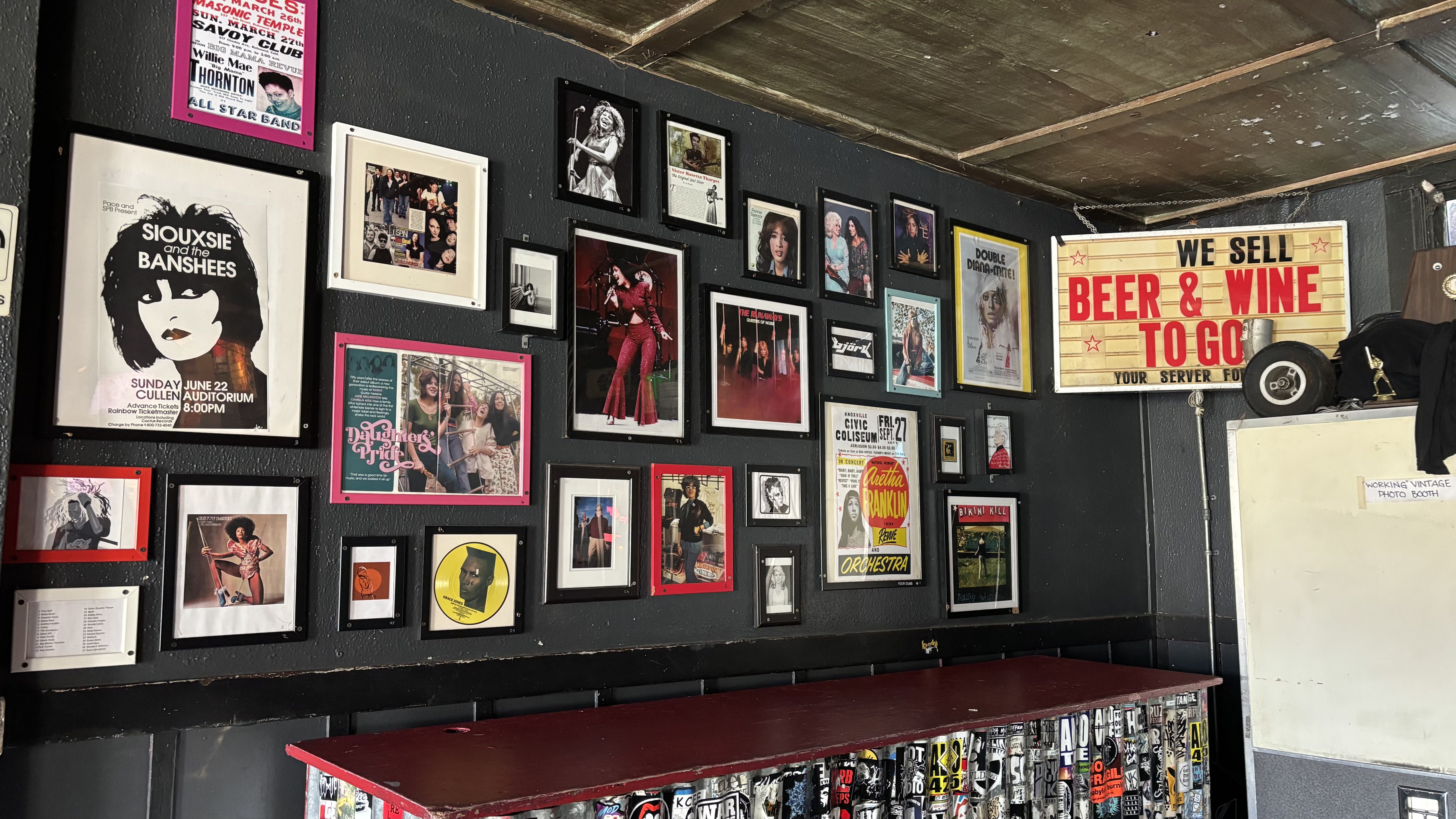 Wall with framed posters and photos of musicians and bands, a red counter below, a sign saying "WE SELL BEER & WINE TO GO" in red and black letters on a white background.