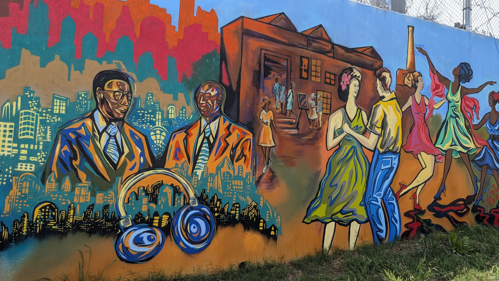 Colorful mural on a wall: a blue cityscape on the left with teal buildings, two men in orange suits, a brown building in the center with people, and dancers in bright dresses on the right under a blue sky.
