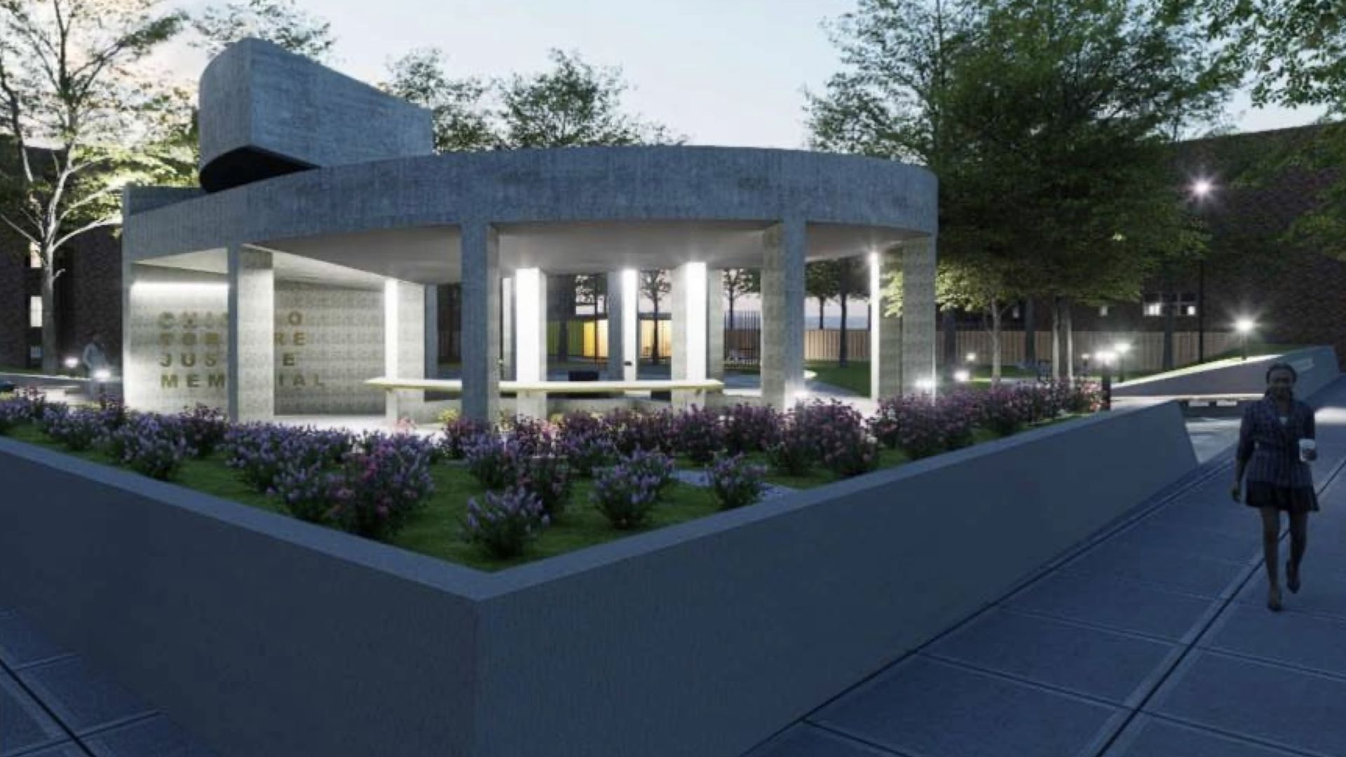 Evening view of a modern circular concrete memorial pavilion in a landscaped park, illuminated by columns, with a low inscribed wall, purple flower beds, trees, and a person walking along a paved path.