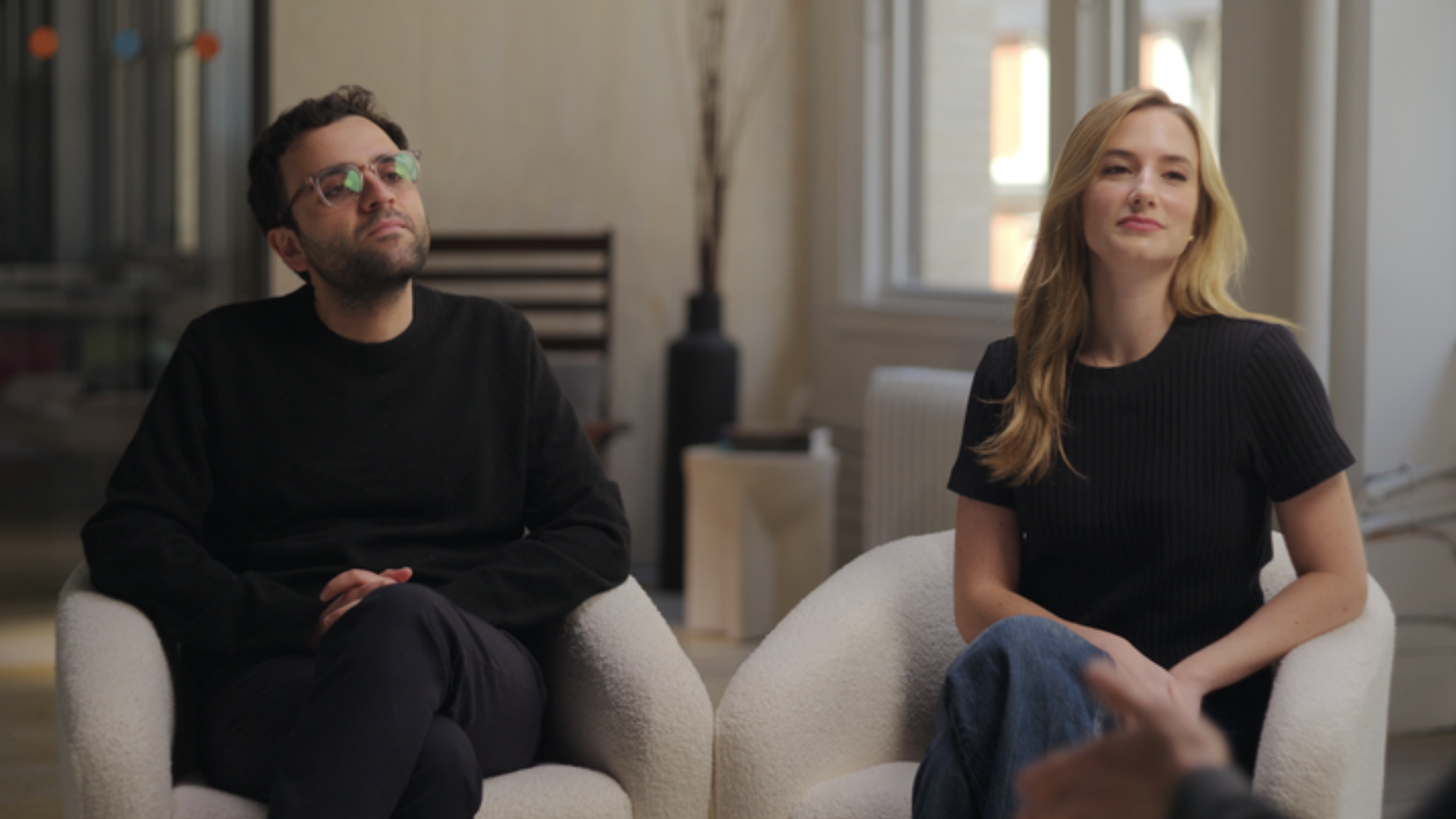 Two people sit in white plush armchairs in a bright, modern room. A bearded man with glasses in a black sweater gazes upward, while a blonde woman in a black top sits beside him, smiling.