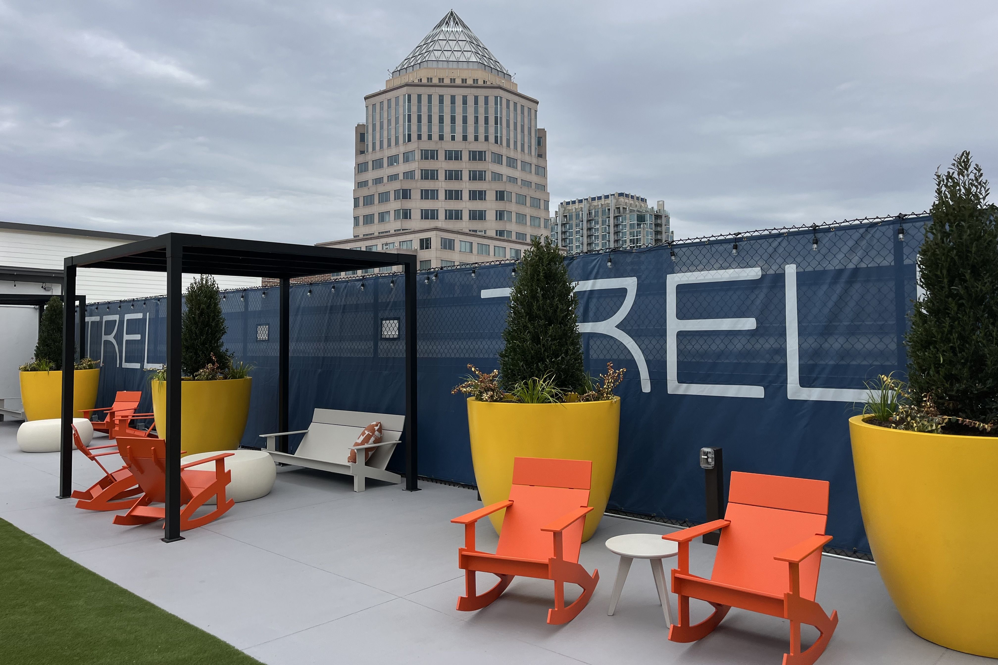 Outdoor seating area with bright orange rocking chairs, large yellow planters with greenery, a gray bench under a black pergola, and tall buildings under a cloudy sky in the background.