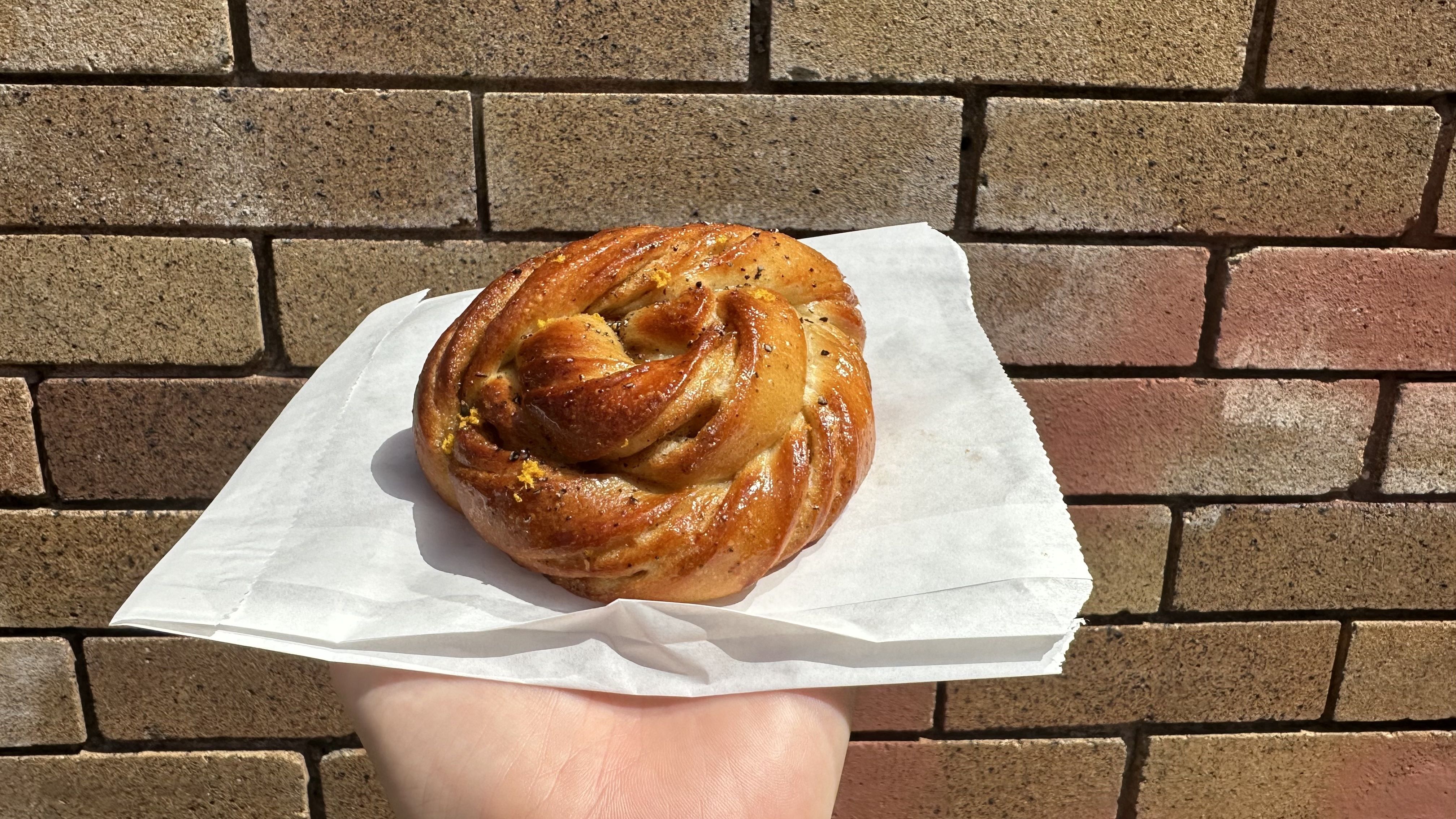 A braided morning bun sits on a white paper as someone holds it up in front of a brick wall.