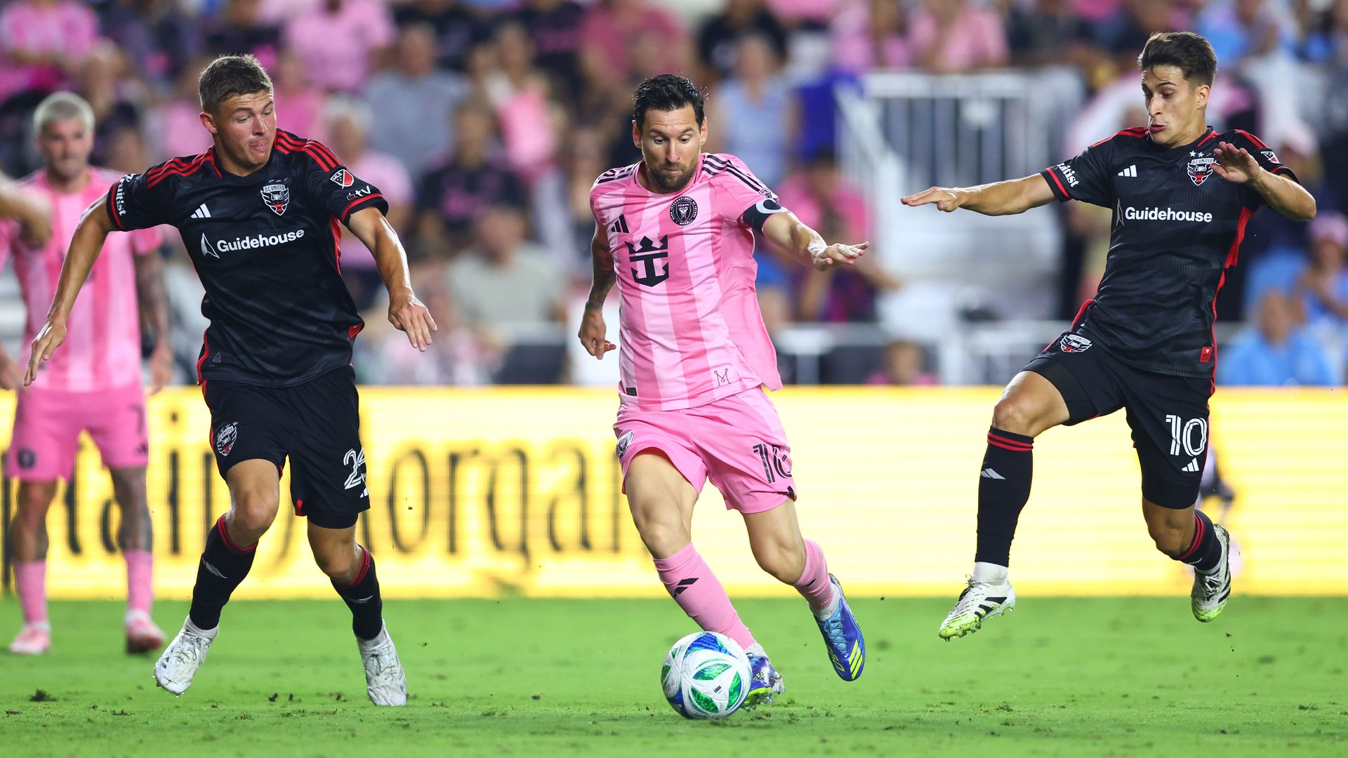 Lionel Messi in pink kit dribbling ball between two D.C. United players in black kits during a match, with an audience in the background wearing pink and other colors.