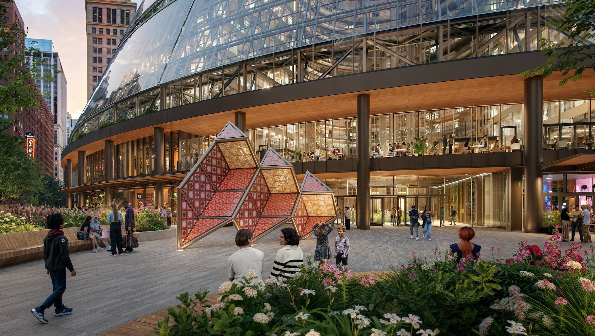Glass-and-steel building with a curved atrium dominates a busy urban plaza. People stroll past flower beds as a large colorful geometric sculpture sits in the foreground.