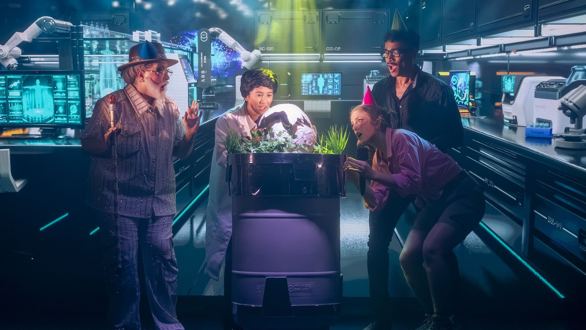 Four performers stand around a puppet display of a hatching dinosaur egg in a lab, in a staged theatre production of Jurassiq Parq.