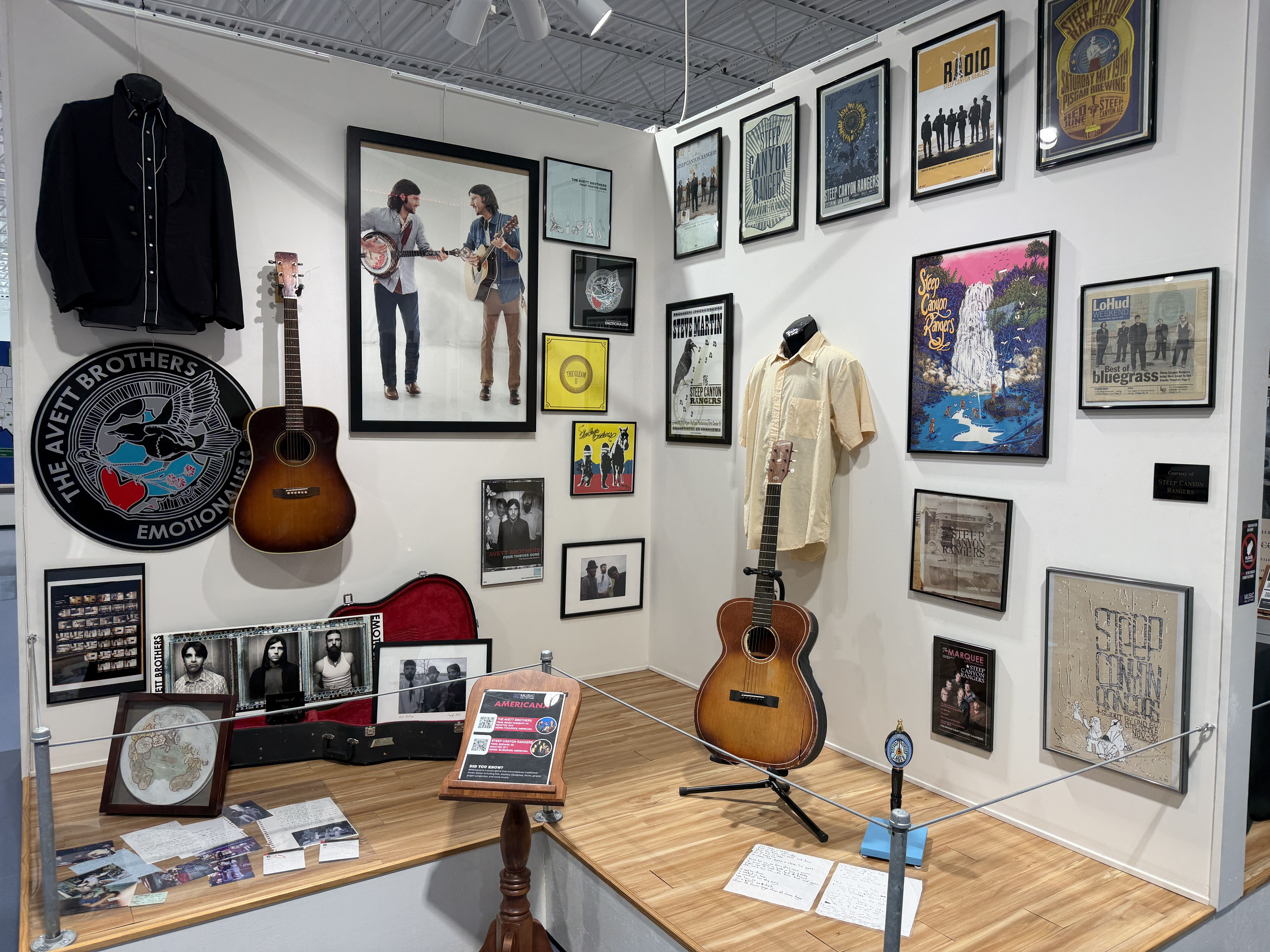 Music exhibit corner: white walls lined with framed posters, two guitars (wall and stand), a beige shirt on a mannequin, and a large circular patch reading "The Avett Brothers".