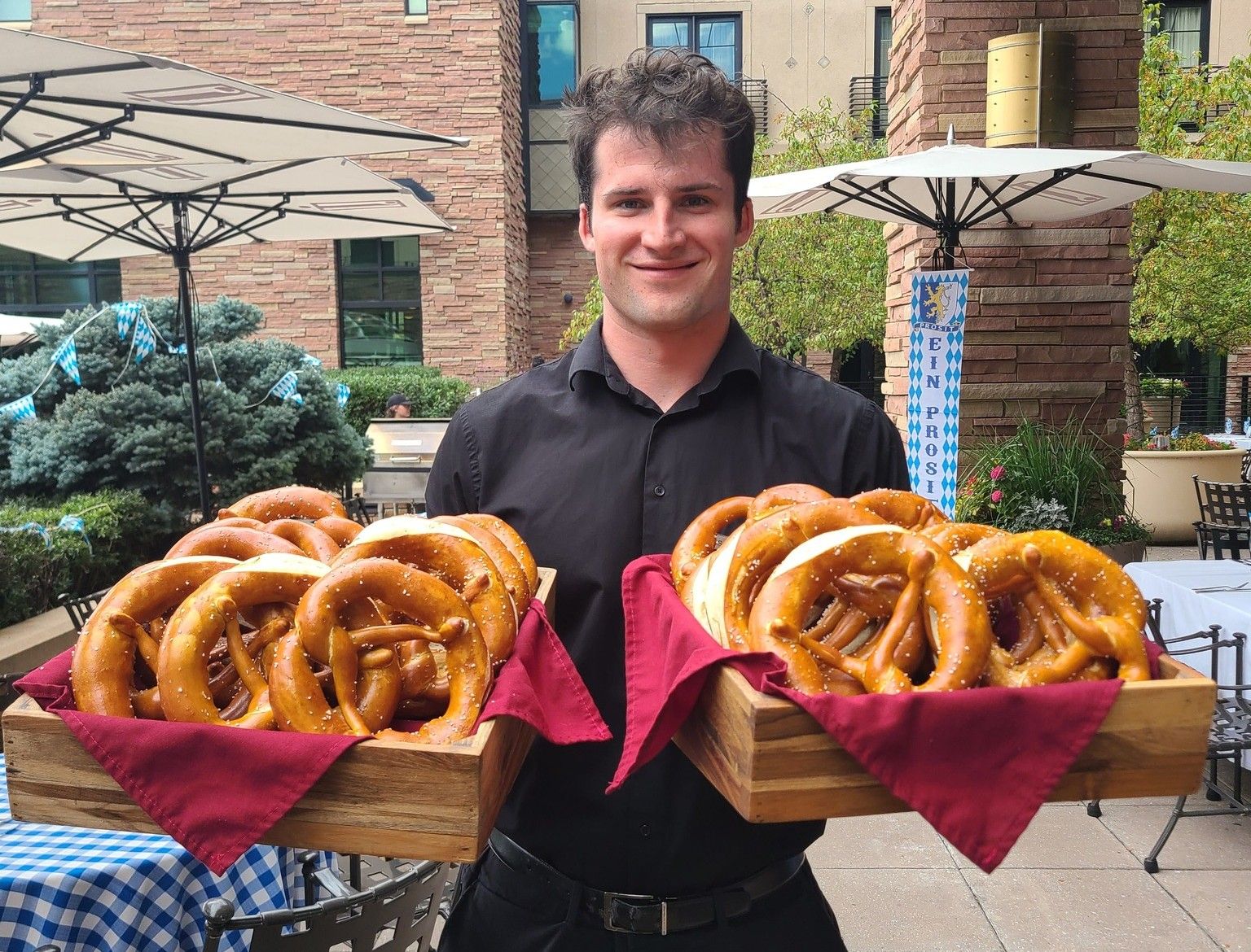 Smiling waiter in a black shirt holds two wooden trays overflowing with large, salted pretzels, lined with red napkins, outdoors under umbrellas at a Bavarian-themed event.
