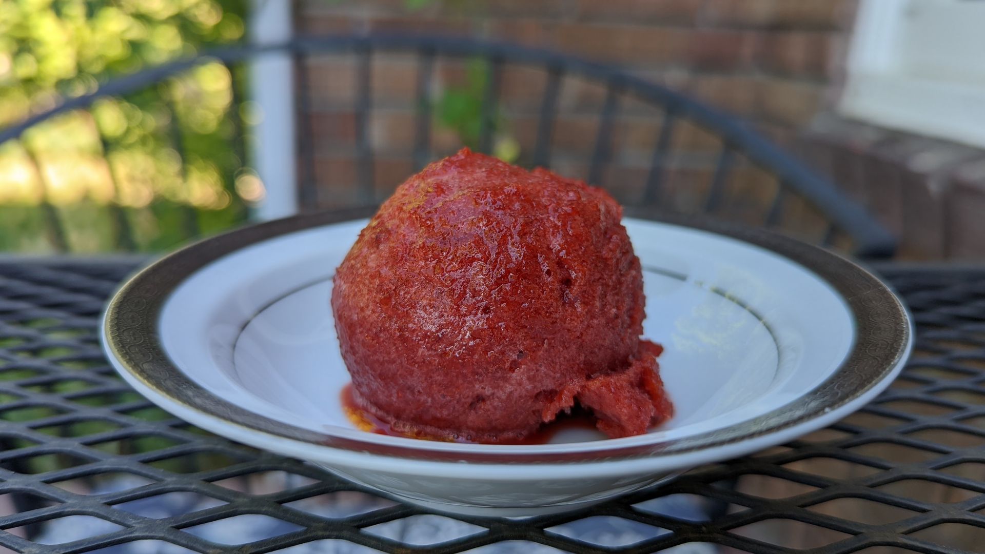 A scoop of sour cherry sorbet in a white bowl on a wrought-iron breakfast table with bricks and plants in the background.