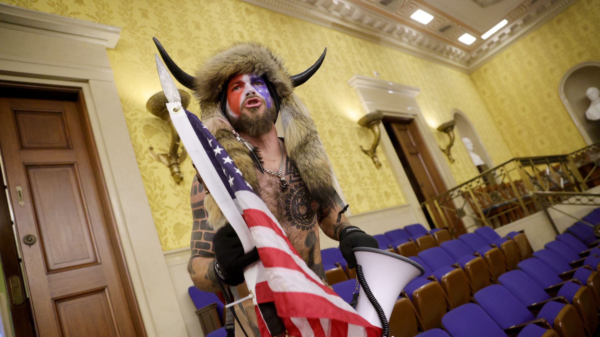 Photo of the QAnon shaman in the Senate chamber holding an American flag and wearing horns