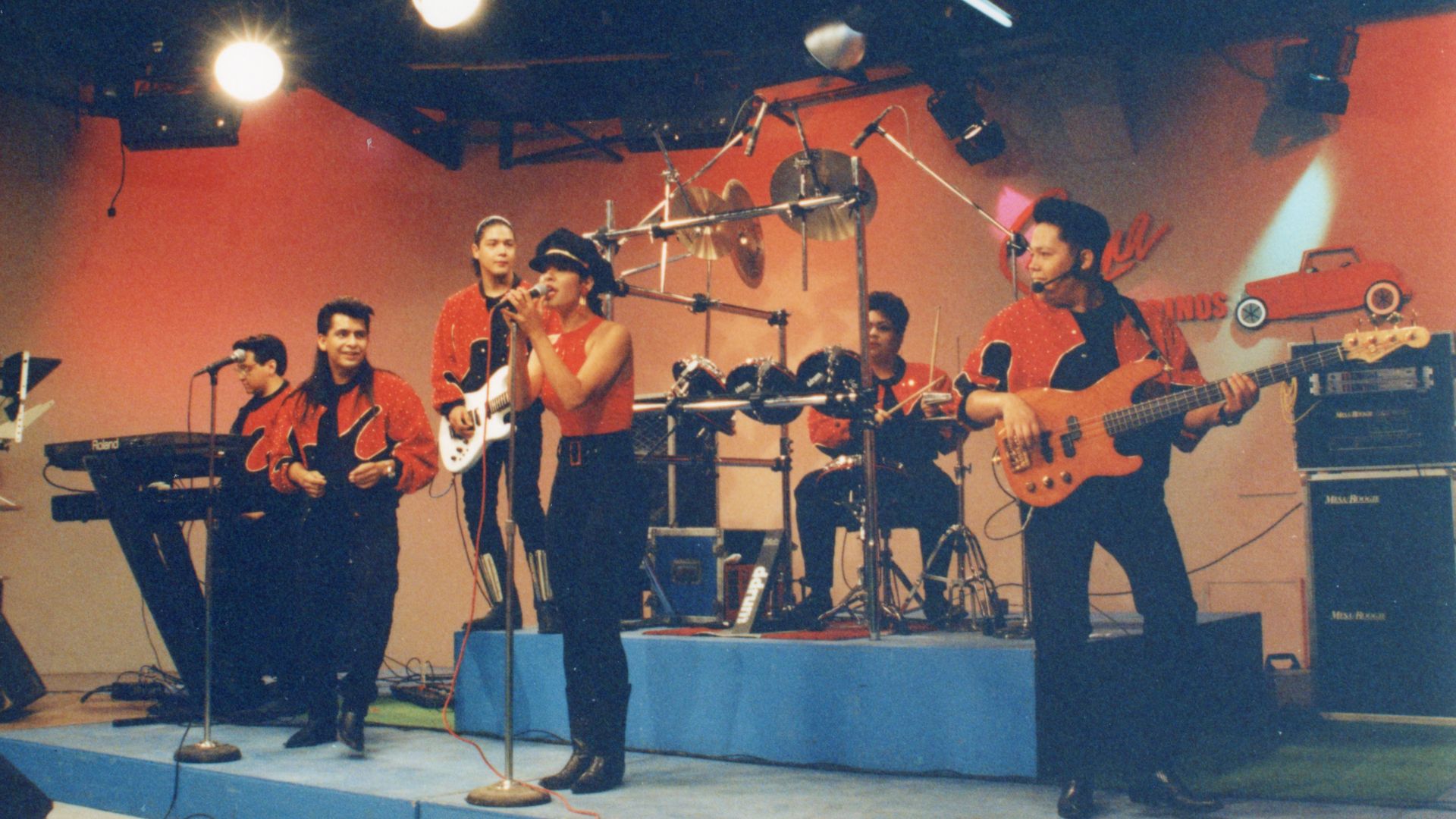 Selena performing on stage with five band members, all wearing red and black outfits.