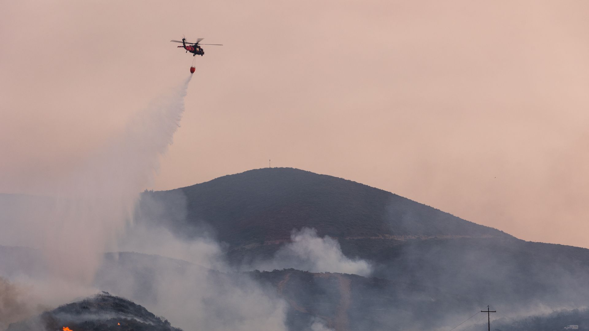 Water falls from a helicopter. The air is smoky and there's a mountain in the background. 