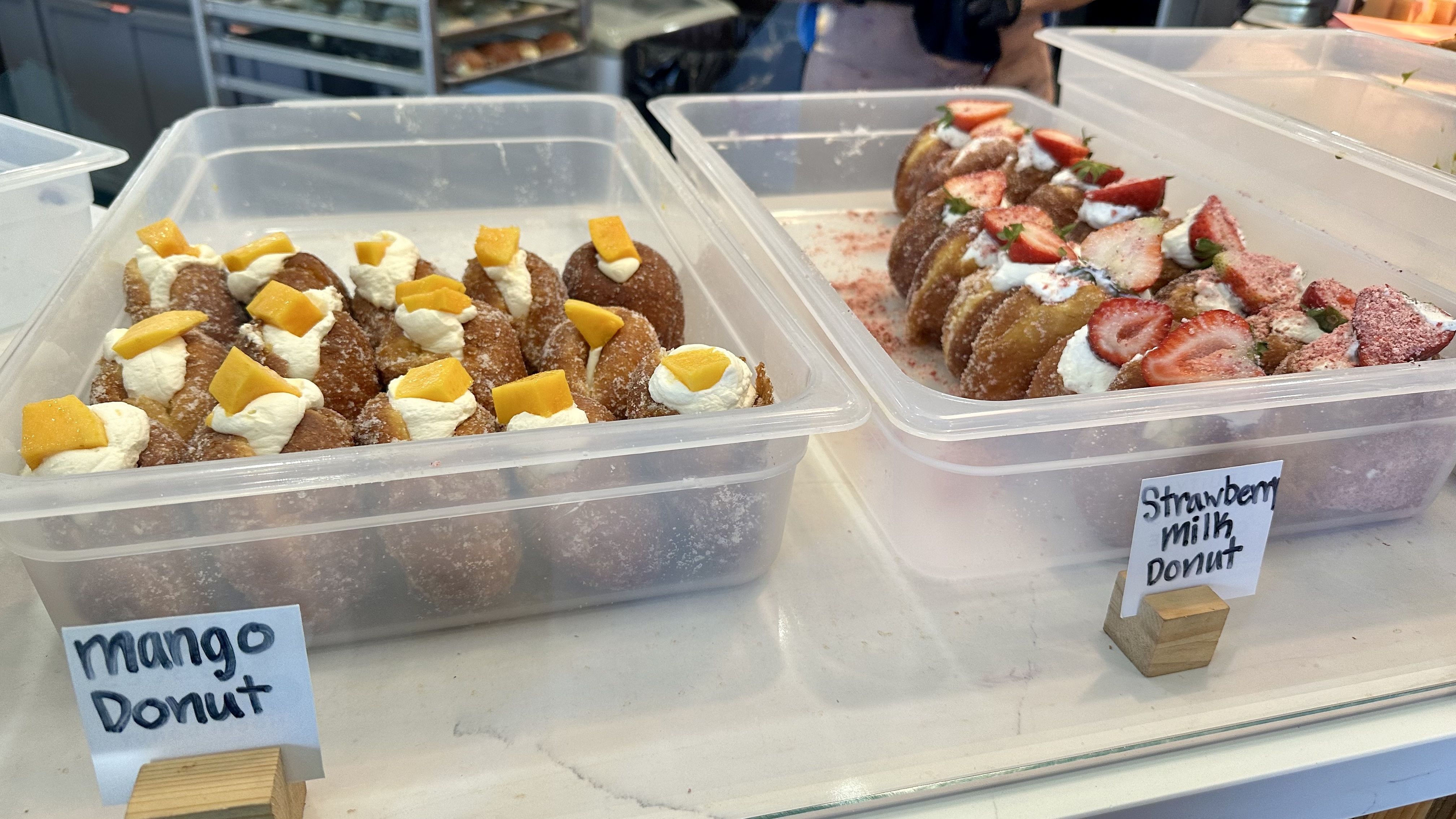 Two clear plastic trays of donuts: left features cream-topped donuts with mango chunks, right features strawberry-topped donuts with cream, strawberries, and pink dust, labeled "Strawberry Milk Donut".