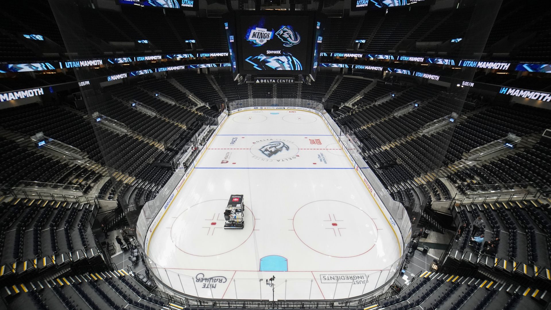 Panoramic view of a large empty hockey arena with dark seating and blue Utah Mammoth banners around the rink, a central scoreboard with logos, and Delta Center signage above the ice.