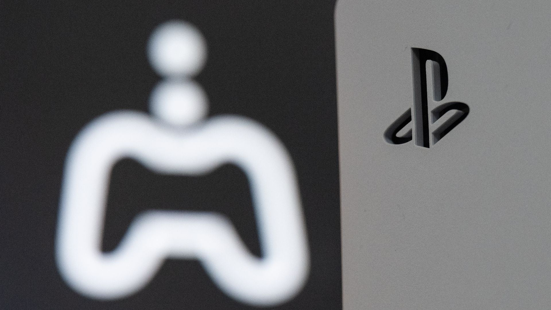 Close-up of a white PlayStation console with black PlayStation logo on the right and a blurry white game controller icon on a dark background on the left.