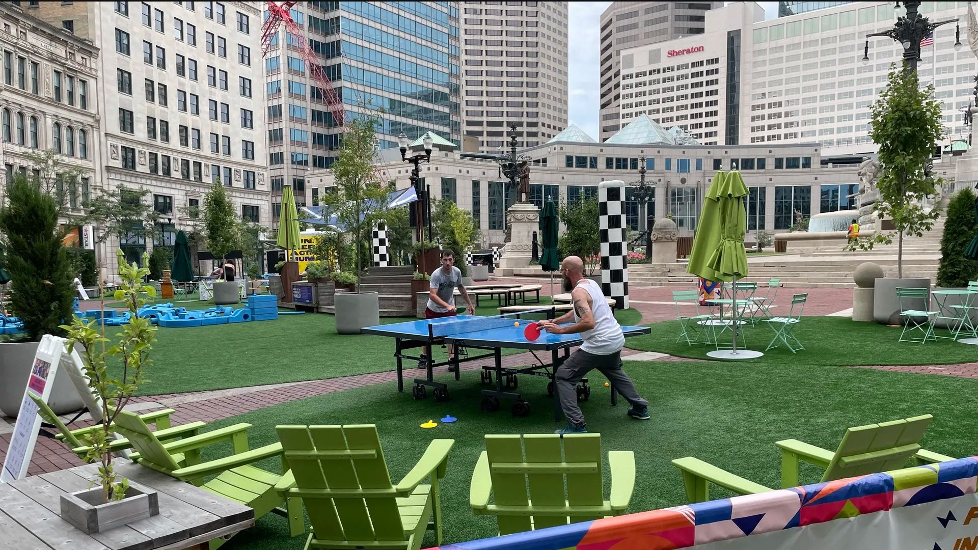 Urban plaza with artificial grass and lime-green chairs, a blue ping-pong table where two men play, surrounded by modern buildings, trees, umbrellas, statues, and a fountain in the background.