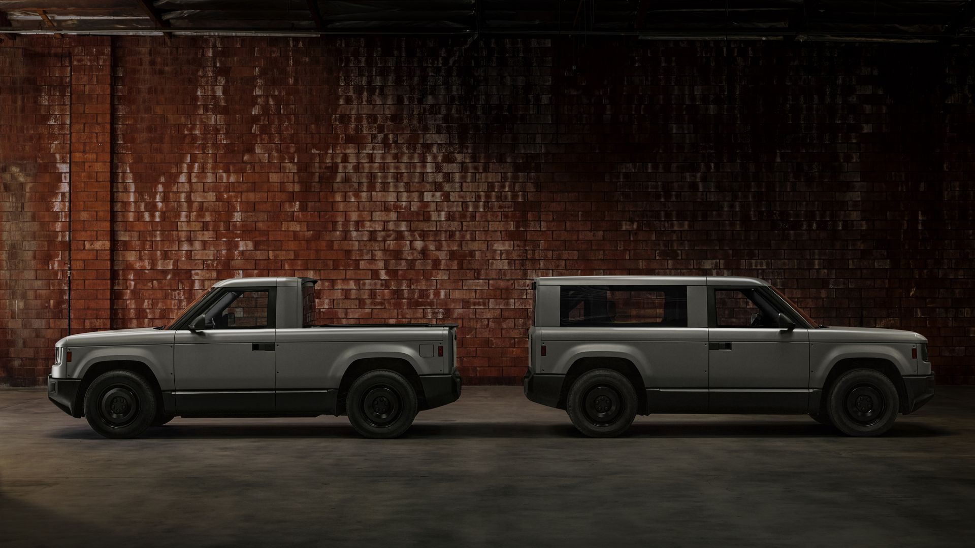Two gray vehicles parked back-to-back in a dim indoor garage with a red brick wall; Left, a Slate pickup; Right, a boxy Slate SUV