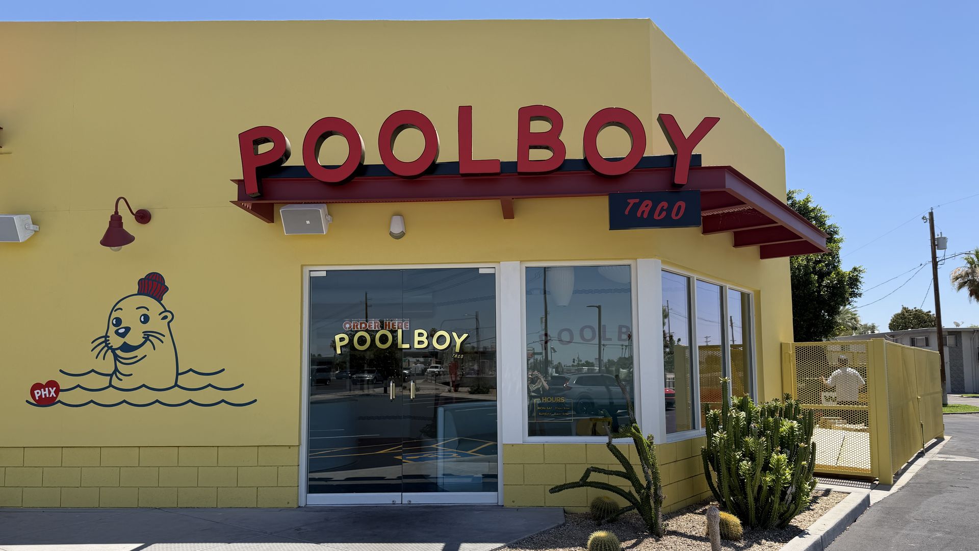Bright yellow fast-food storefront with a large red sign reading "POOLBOY" across the front and a smaller blue sign reading "TACO". A cartoon seal in a chef hat sits on the wall to the left, blue sky overhead.