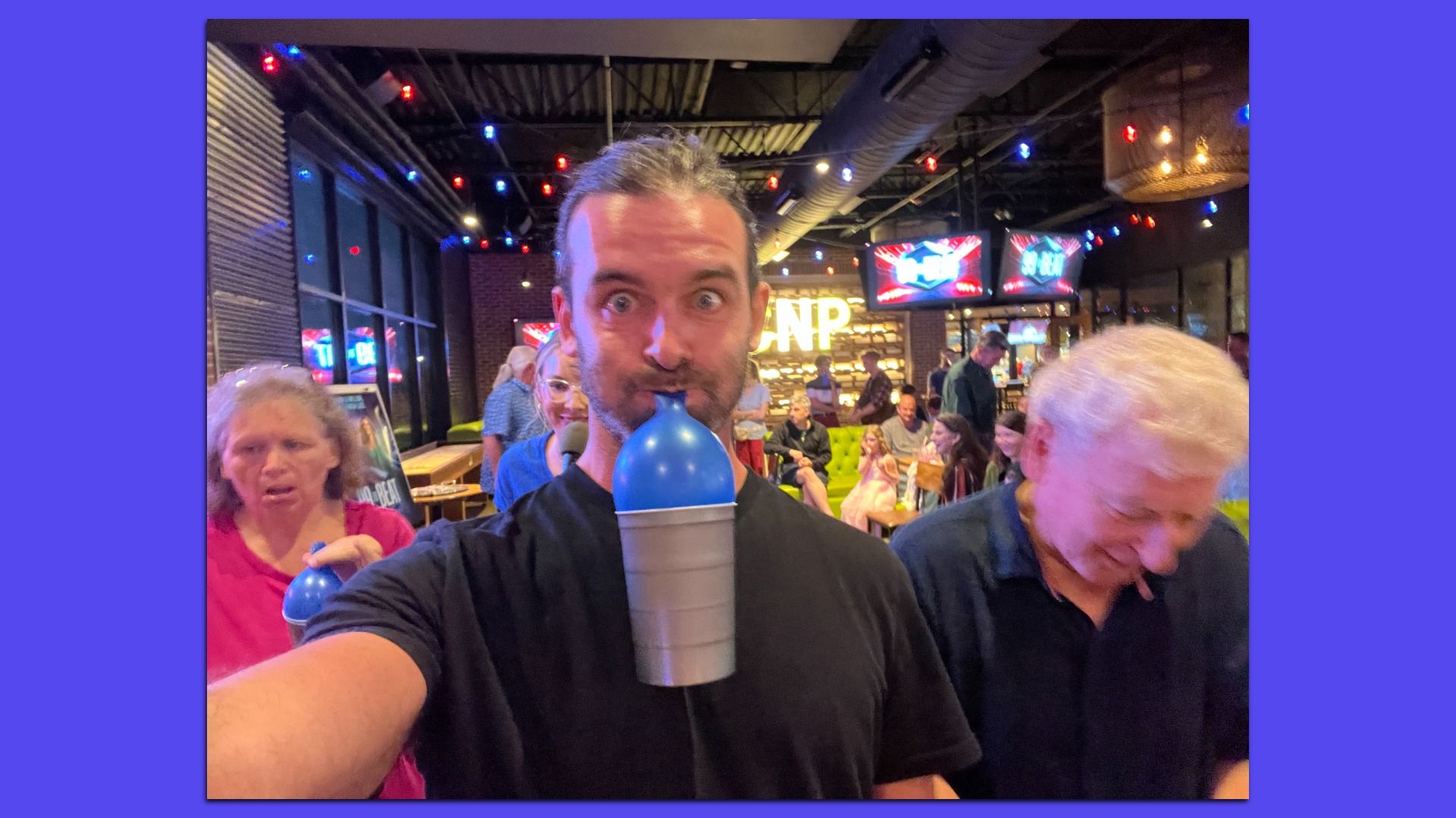 Reporter Travis holding a silver cup with a blue balloon blown up inside hanging from his mouth, indoors with colorful string lights and people socializing in the background at a lively venue.