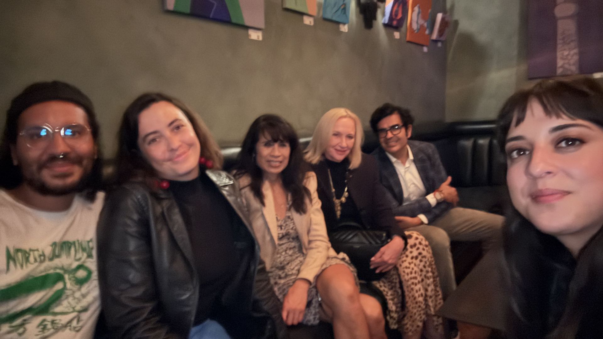 Six friends sit on a dark sofa in a dim room, smiling at the camera. A woman on the right takes the selfie; colorful panels hang on a gray wall behind them.