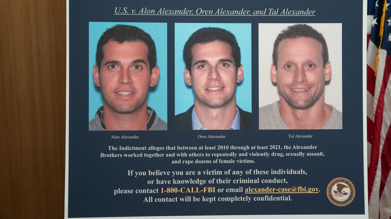 Photo: Axios Poster board with three mugshots on blue panels, a large indictment text block, and an American flag visible to the right