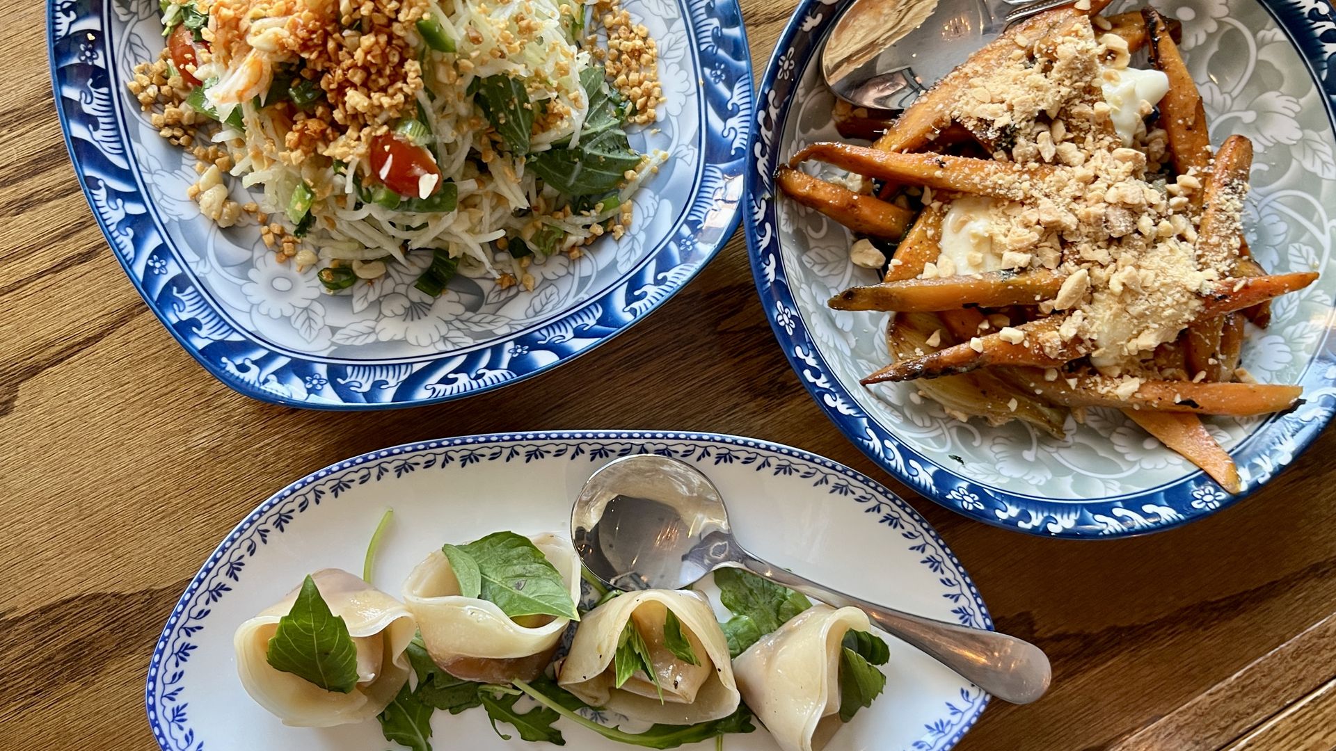 Three blue and white patterned plates on a wooden table with a shredded vegetable salad topped with chopped nuts, roasted carrot sticks with creamy sauce and nuts, and dumplings garnished with fresh greens.