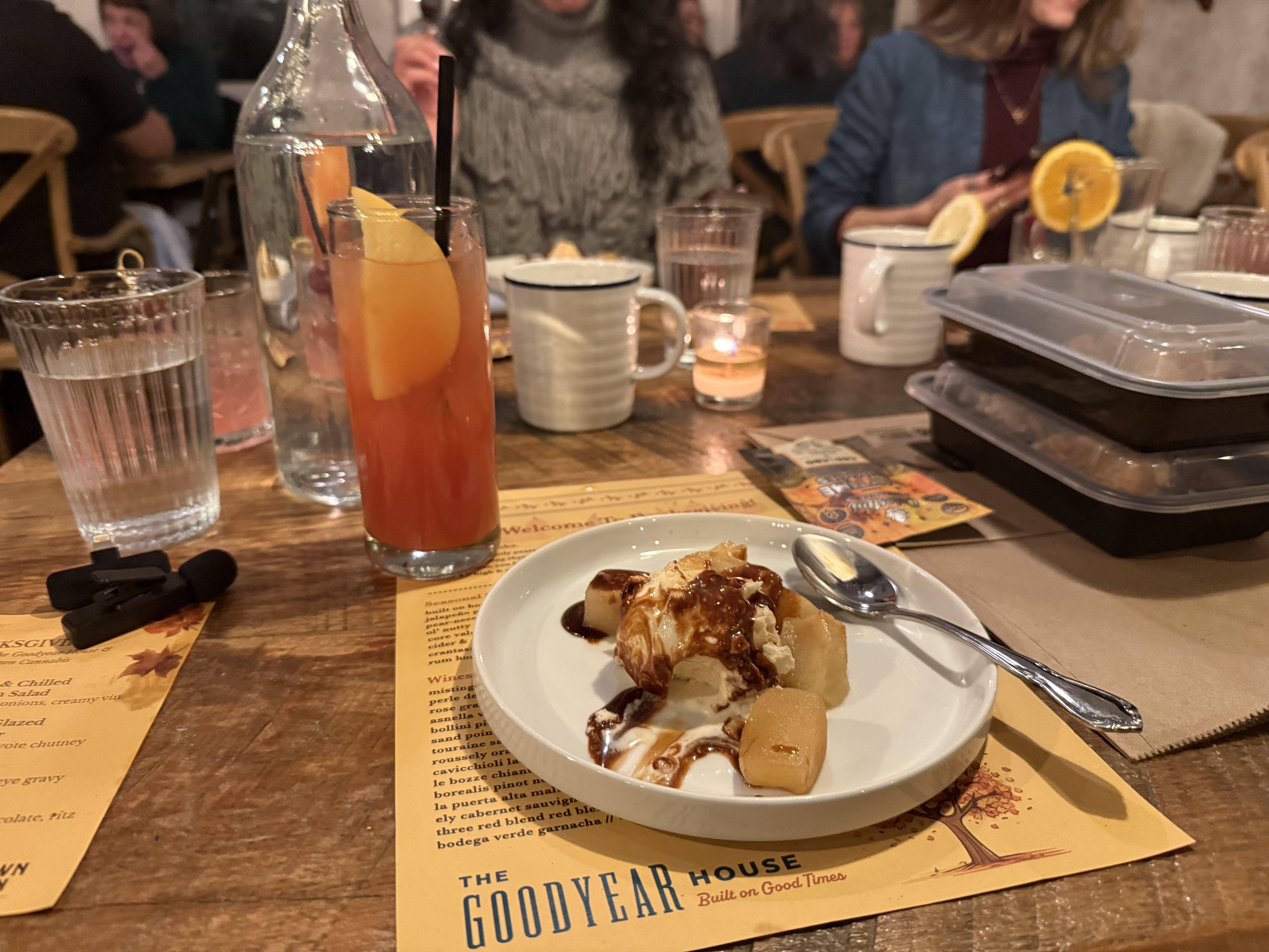 Table with dessert plate of ice cream and caramelized fruit, a tall reddish drink with an orange slice, water glasses, and menus at a restaurant named The Goodyear House.