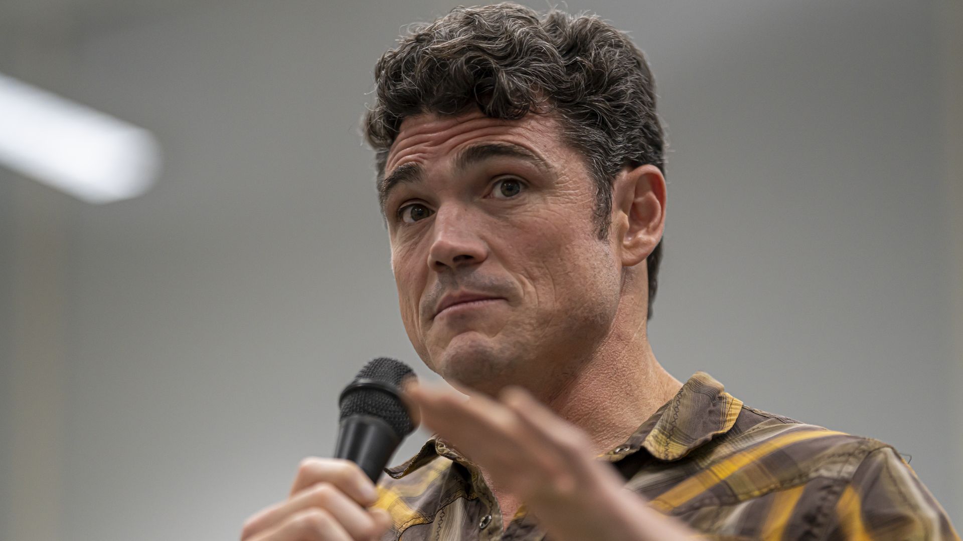 Joe Kent, wearing a brown and yellow button down shirt, holding a microphone in a white room.