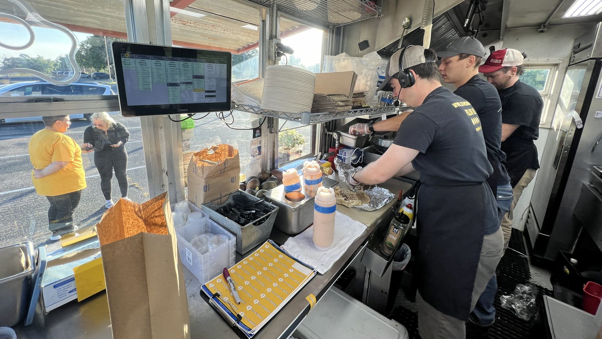 The interior of a food truck with metal kitchen counters, a crew of three men in black shirts and hats preparing food, and a cashier in a yellow shirt visible through the window taking an order from a customer.