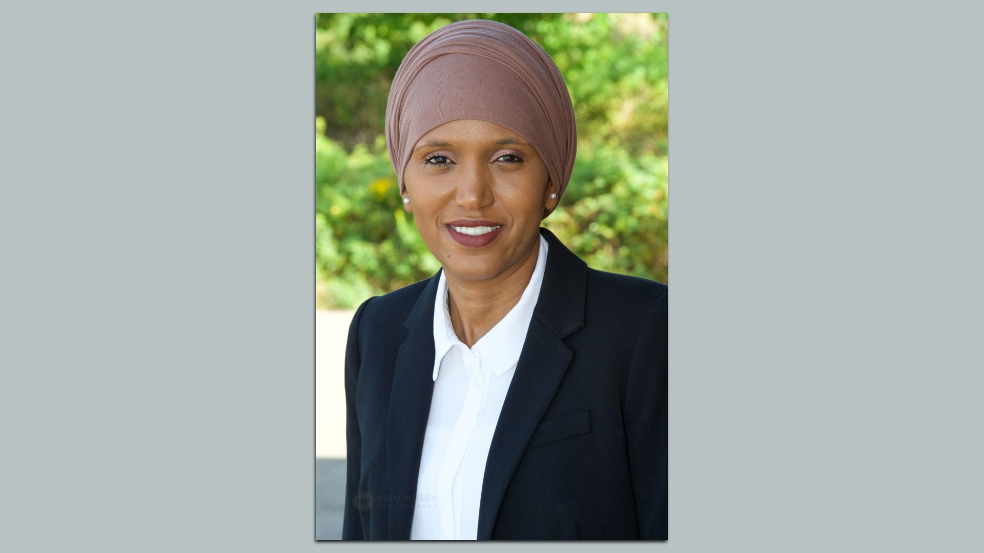 Portrait of a smiling woman wearing a taupe hijab, white blouse, and black blazer, outdoors with green foliage in the blurred background.