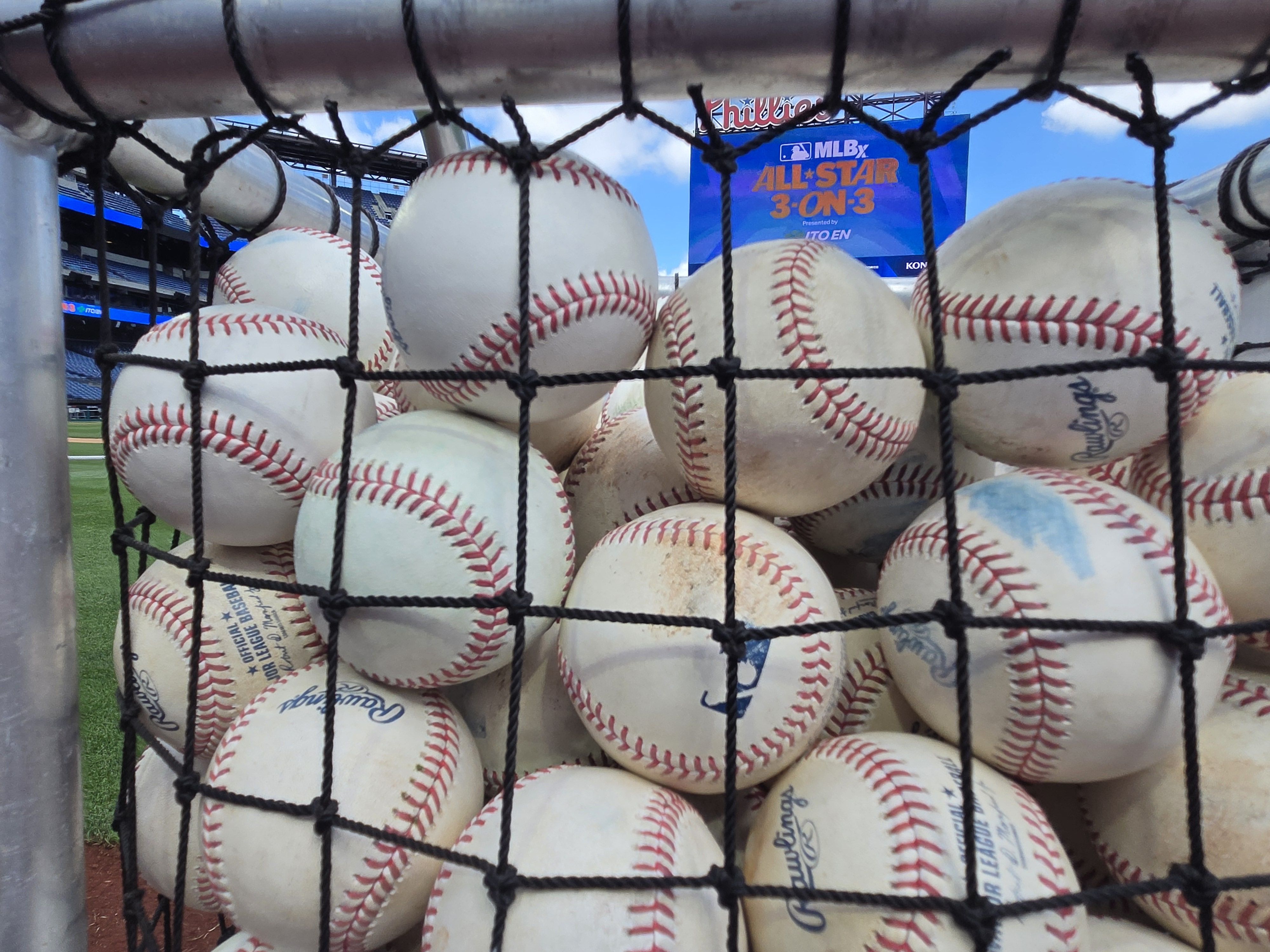Close view of numerous white baseballs with red stitching piled behind a black net, at a baseball stadium. A blue scoreboard in the background shows MLB All-Star 3-On-3 under a bright sky.