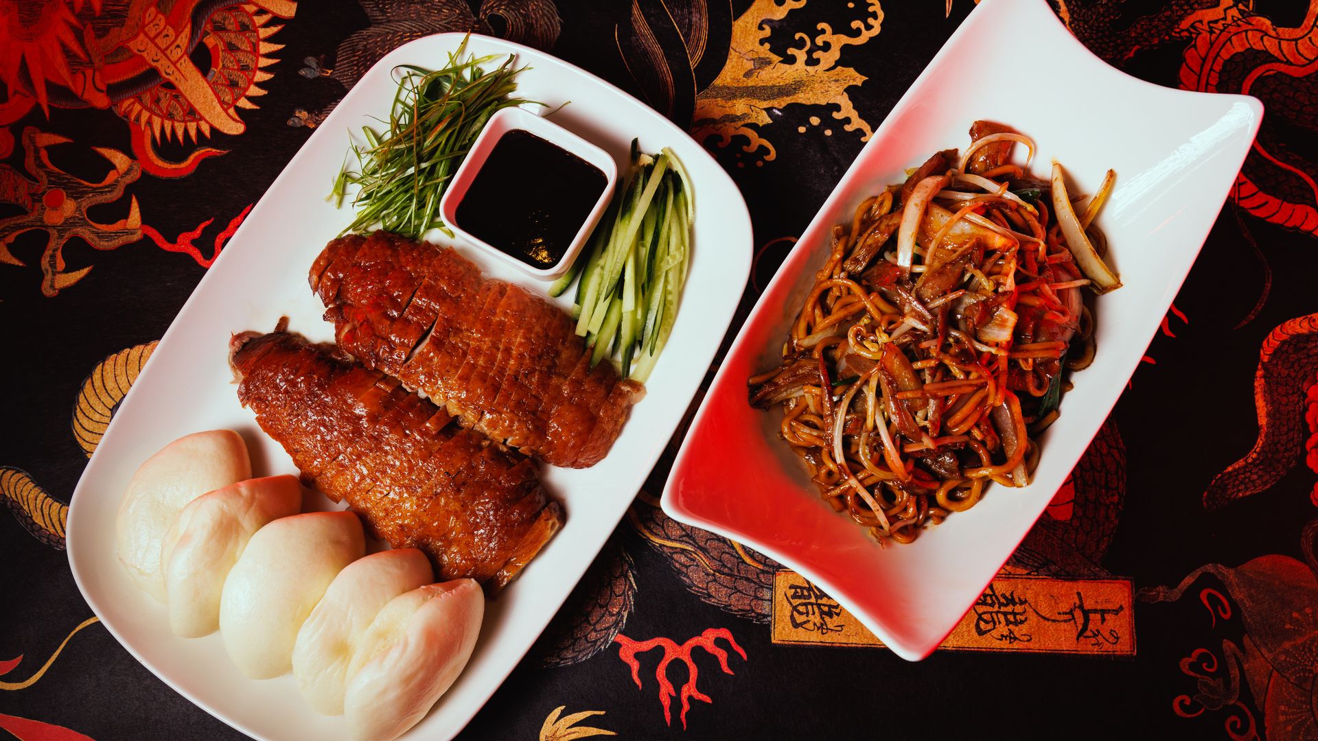 Two white plates on a dragon-patterned tablecloth; one with sliced roasted duck, steamed buns, cucumber, scallions, and dark dipping sauce, the other with stir-fried noodles and vegetables.