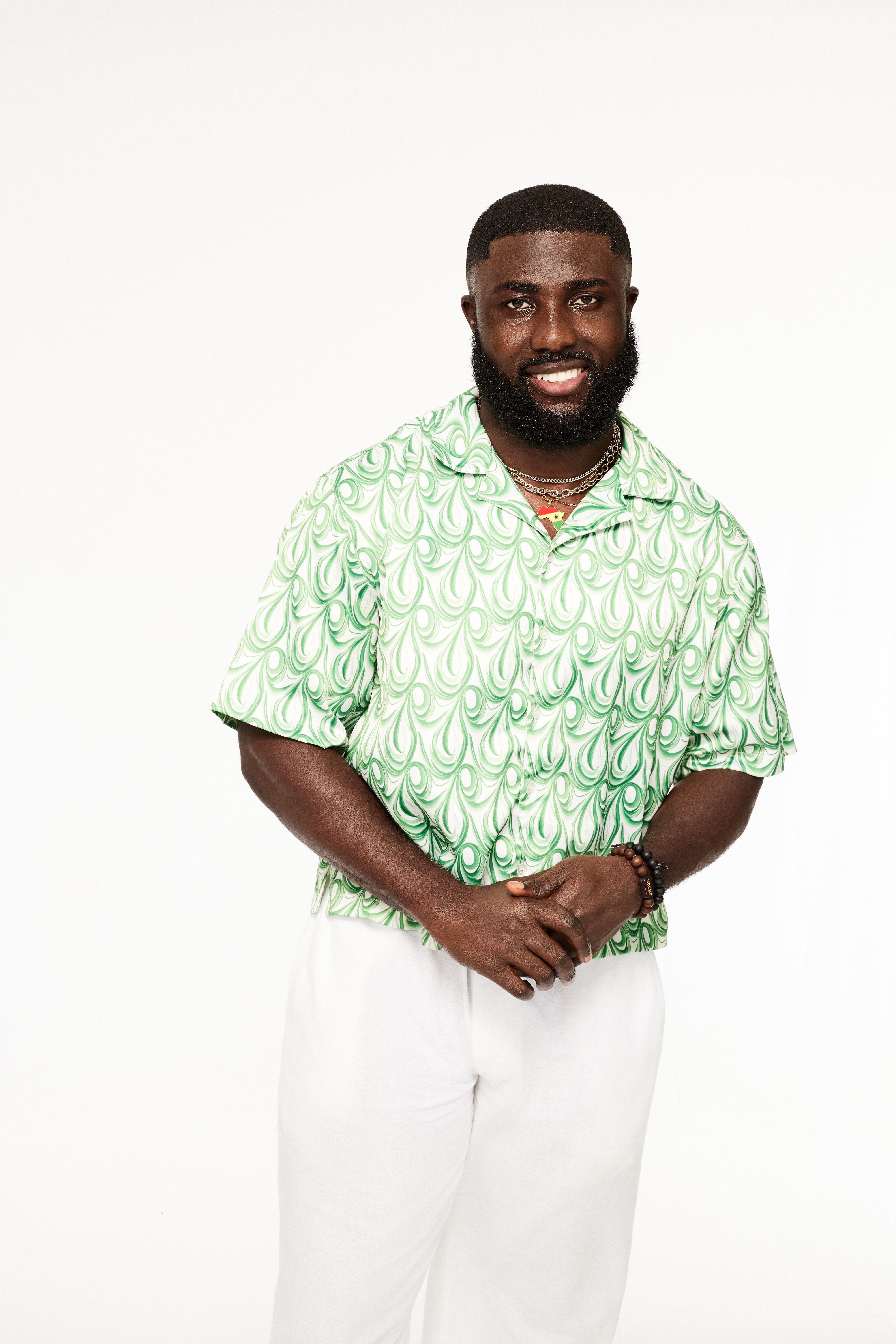 Smiling man with a beard wearing a green and white patterned short-sleeve shirt and white pants, standing against a plain white background with hands clasped.