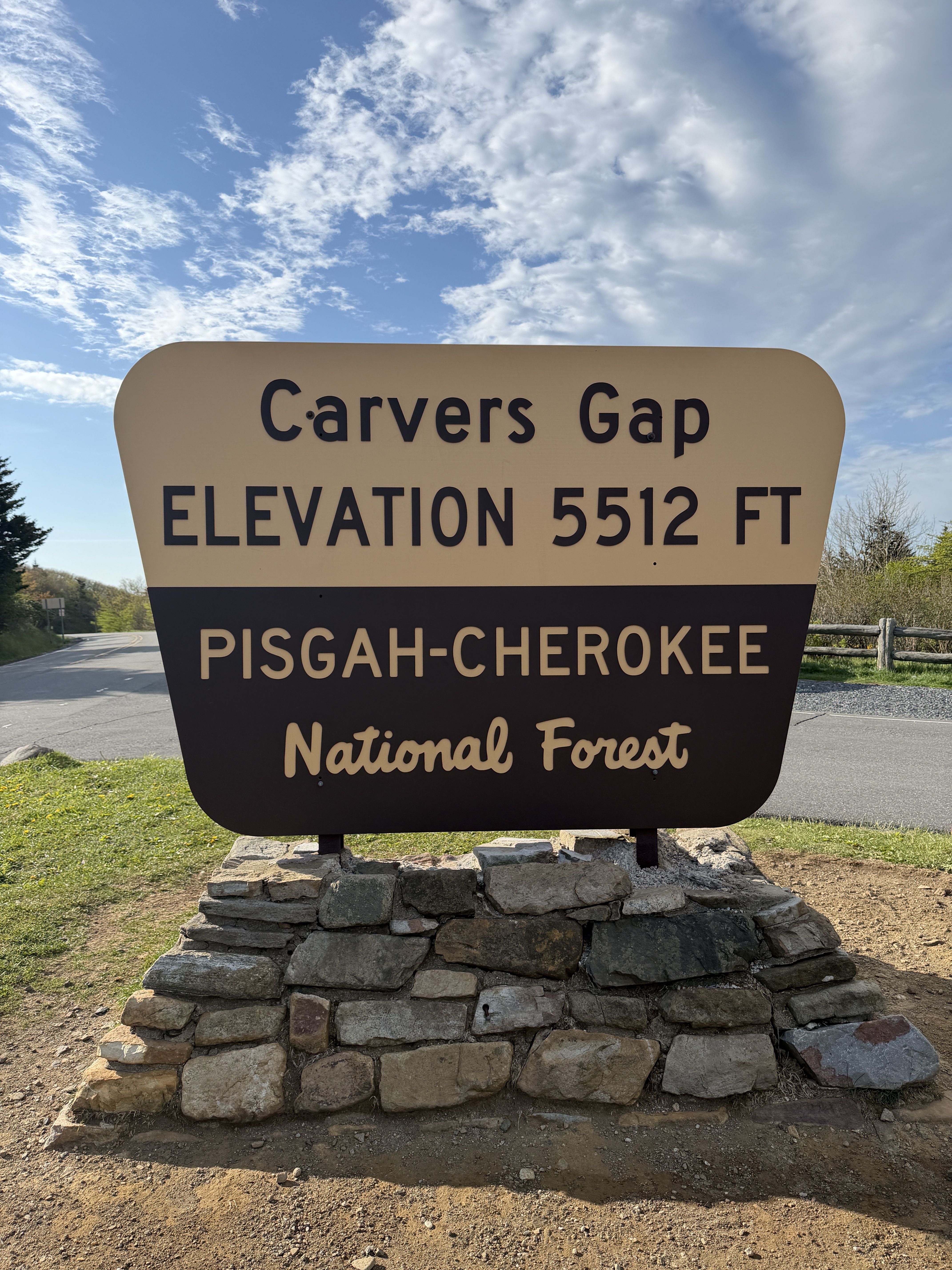 Large rounded roadside sign at Carvers Gap showing elevation 5512 ft, mounted on a stacked stone base; below reads Pisgah-Cherokee National Forest. Blue sky with clouds behind.