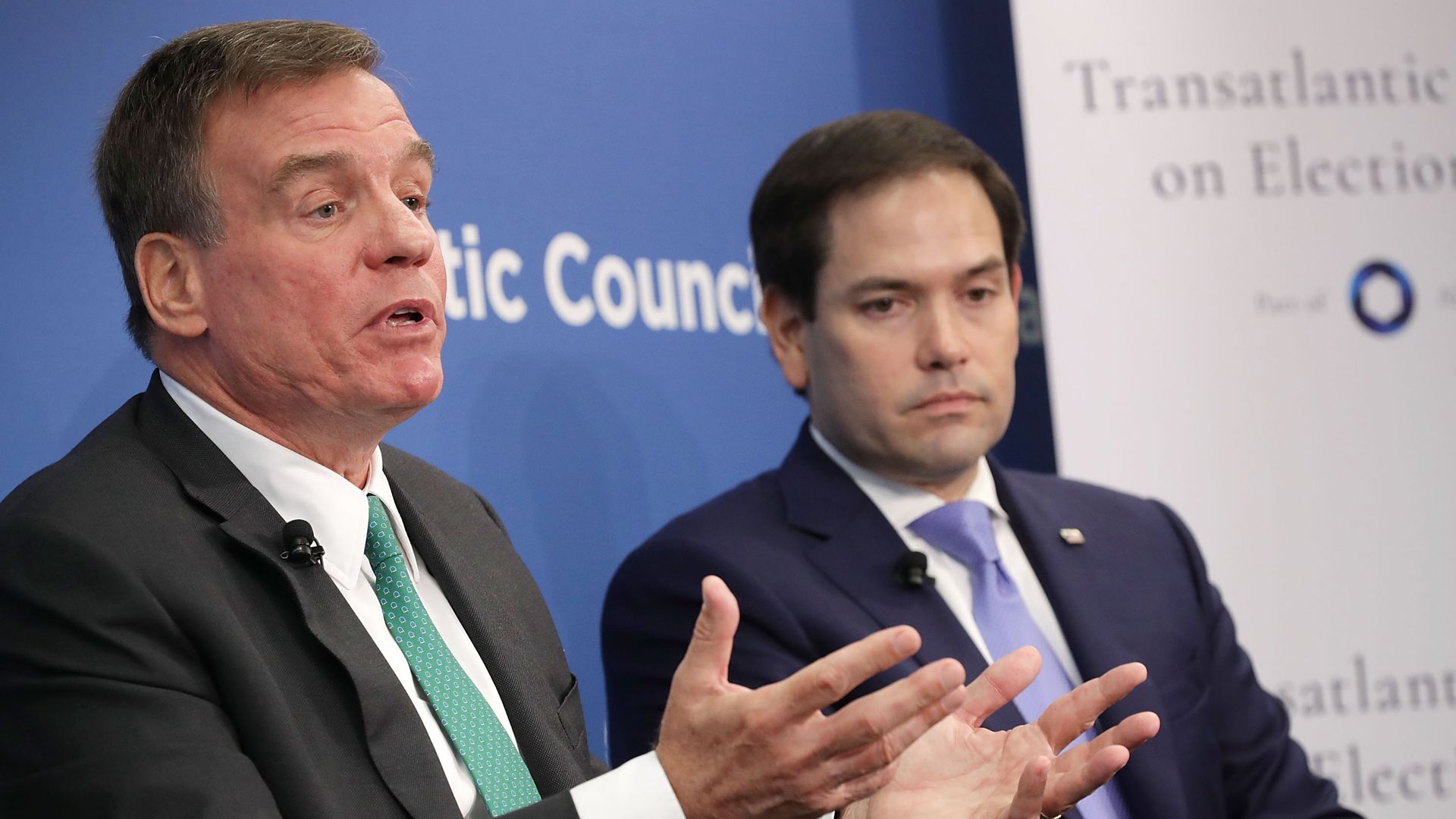 Mark Warner gestures while talking and Marco Rubio looks on sternly