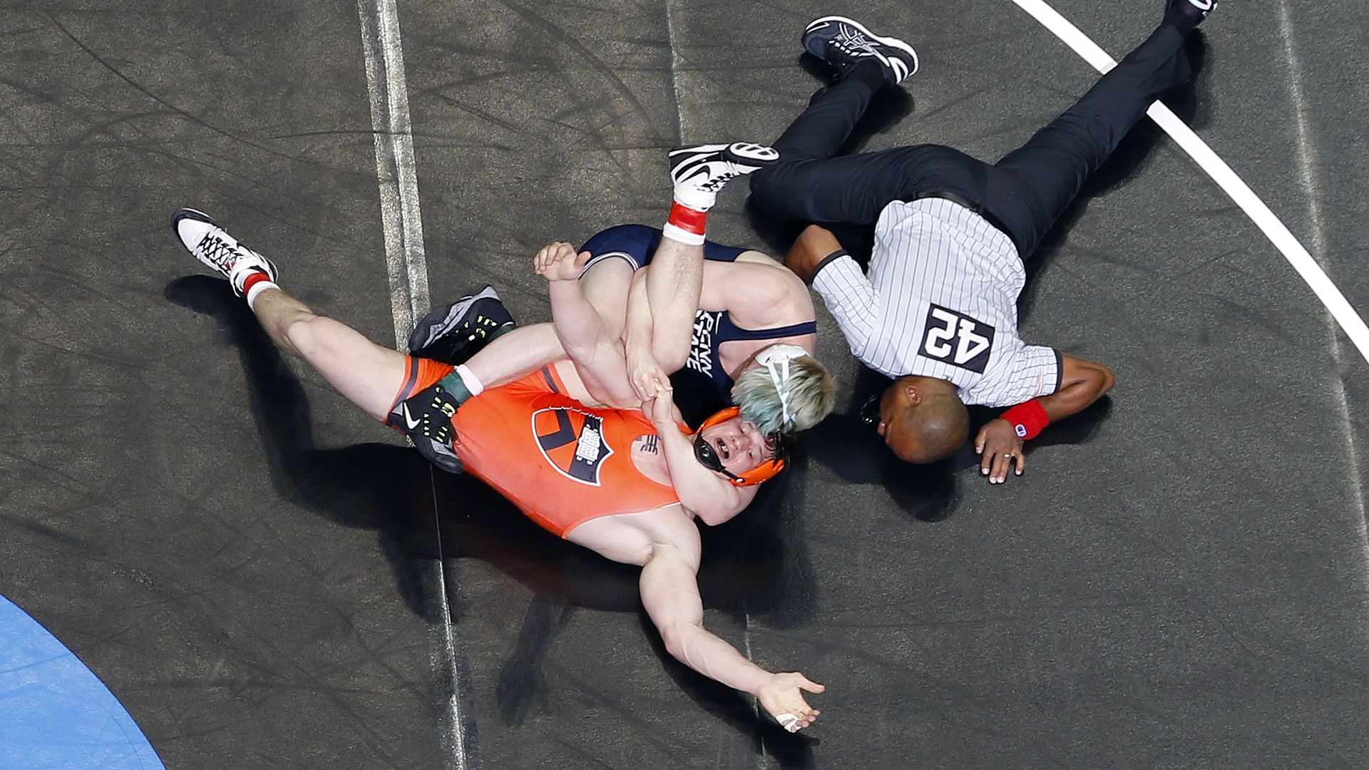 Two wrestlers on a black mat, one in orange and one in dark blue, grappling closely with a referee in a striped shirt and black pants kneeling nearby.