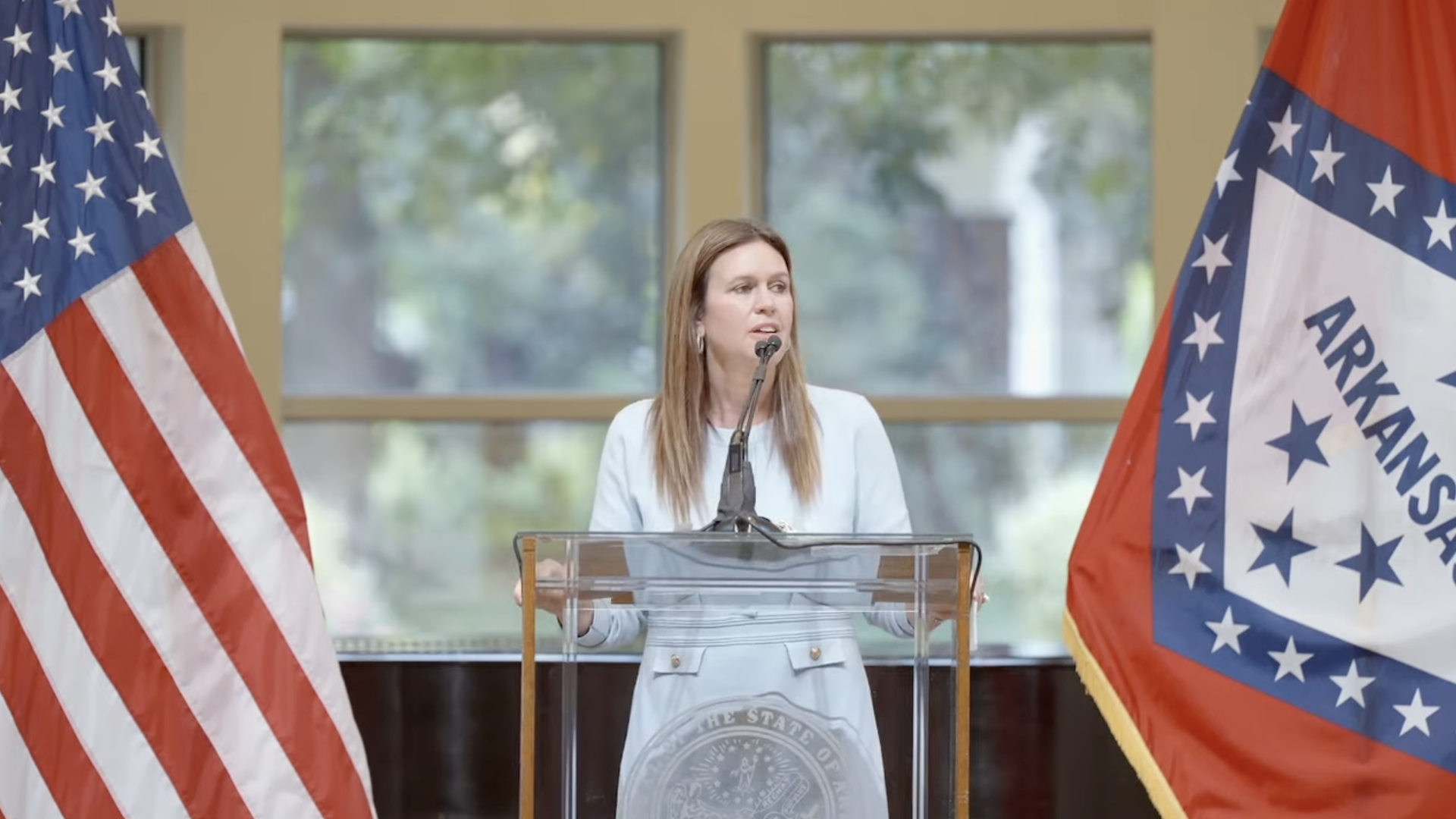 Arkansas Governor Sarah Huckabee Sanders speaks at a podium between U.S. and Arkansas state flags, inside a bright room with tall windows.