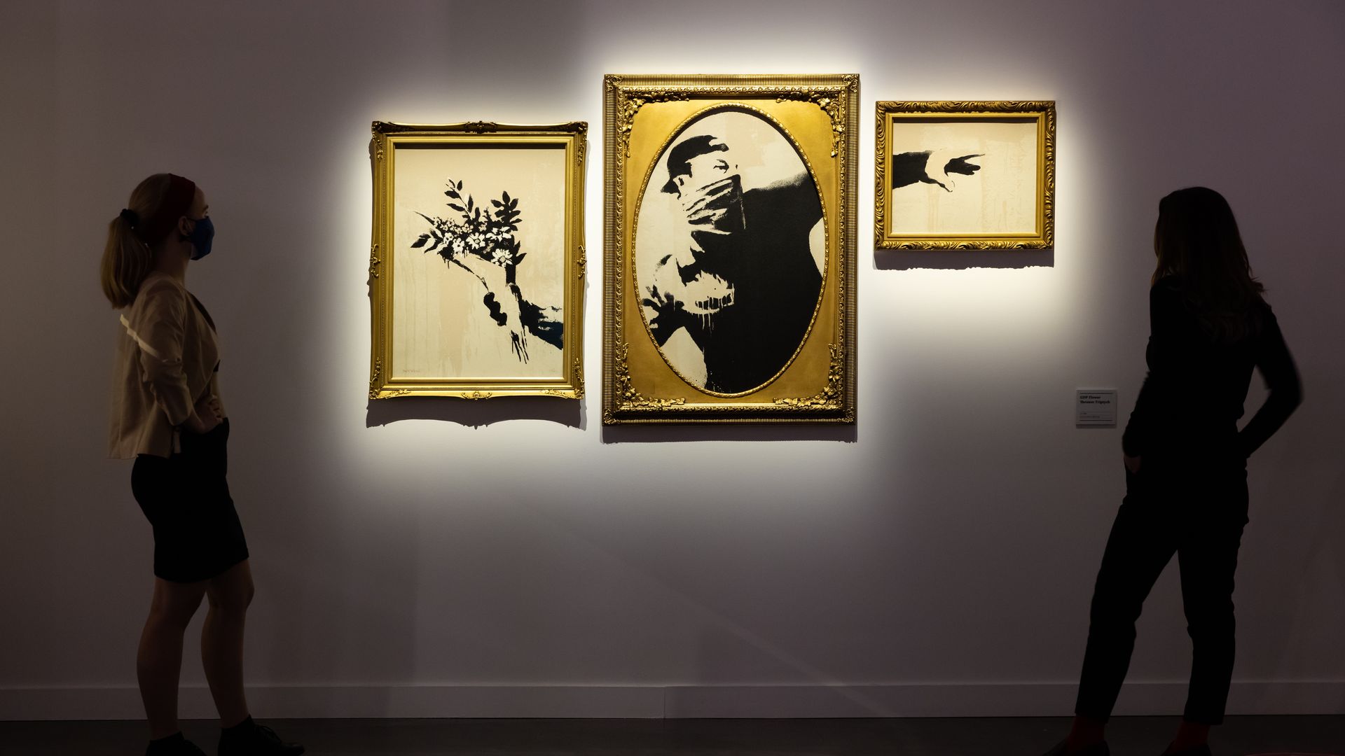 Three pieces of art by Banksy hang on the wall as people gather around. 