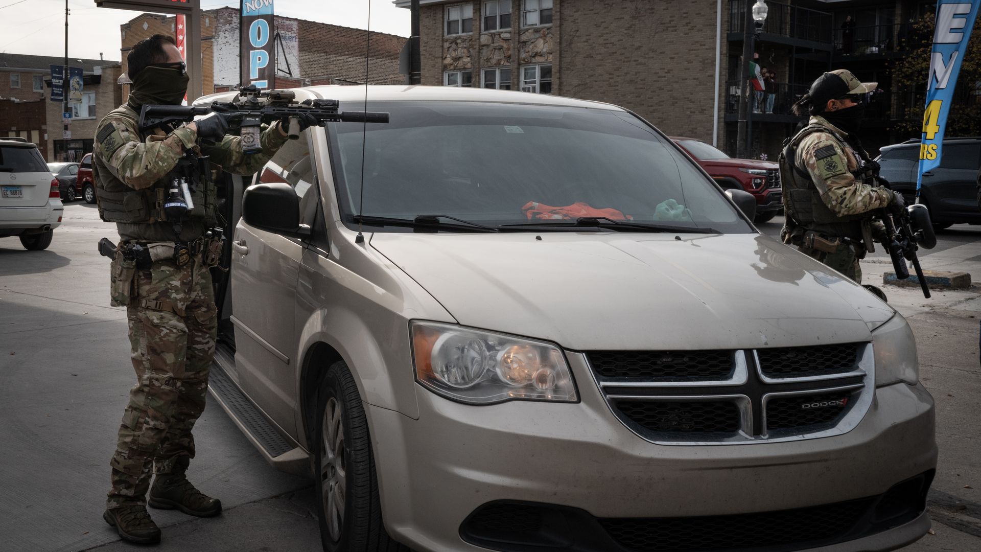 Two heavily armed soldiers in camouflage uniforms stand by a beige Dodge minivan on a city street, wearing masks; urban buildings and a gas price sign in the background.