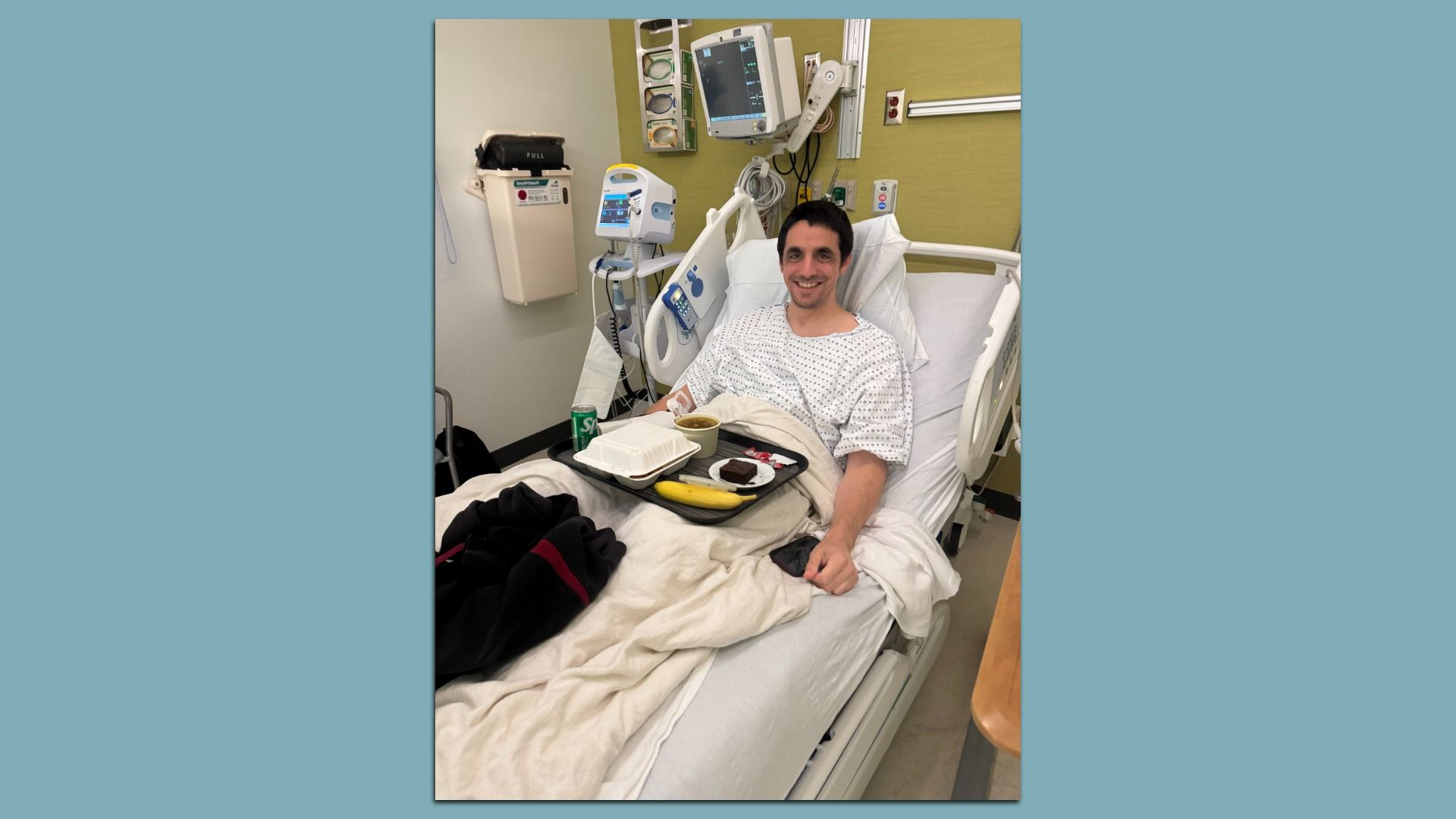Smiling man in hospital gown lies in bed with beige blanket, hospital monitors behind him, and a tray with food including a banana, brownie, soup, and soda can on his lap.