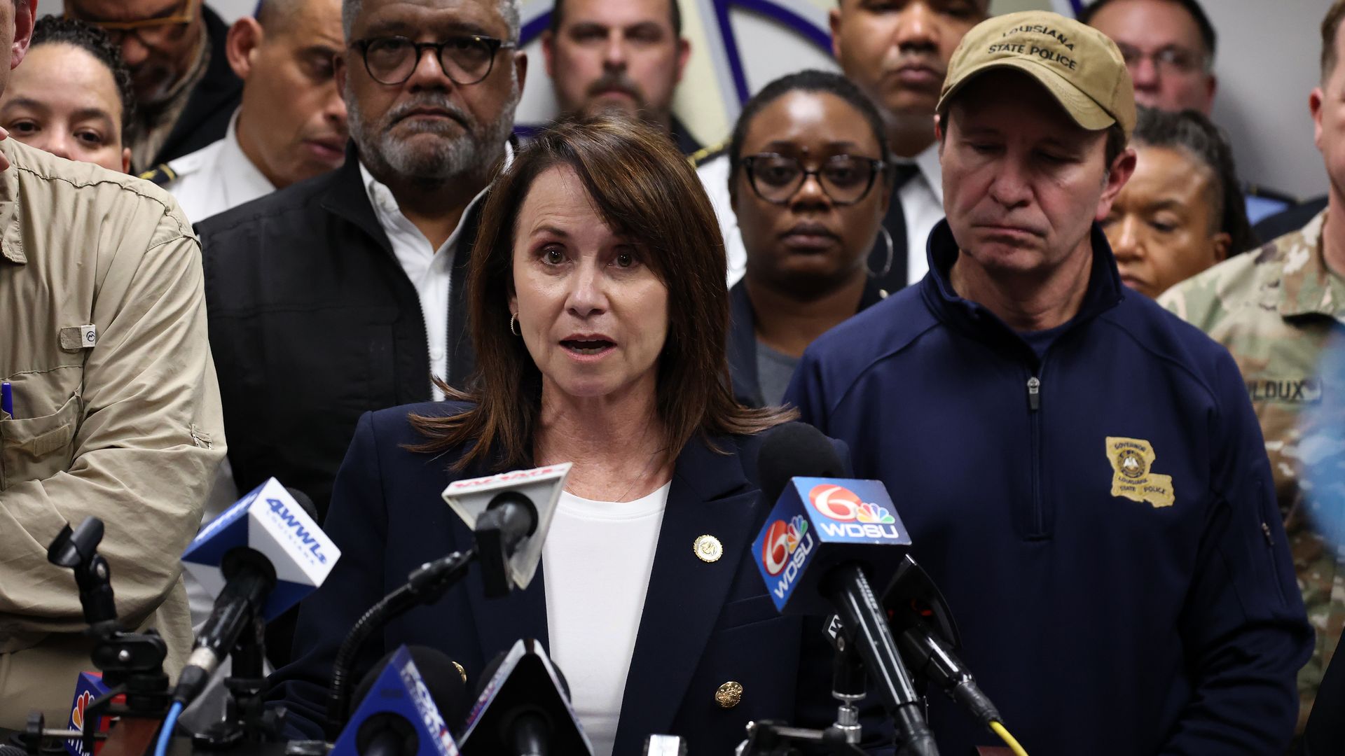 Liz Murril speaks at a podium with several microphones clipped to its edge. Behind her is a crowd of people, including Gov. Jeff Landry looking on. 