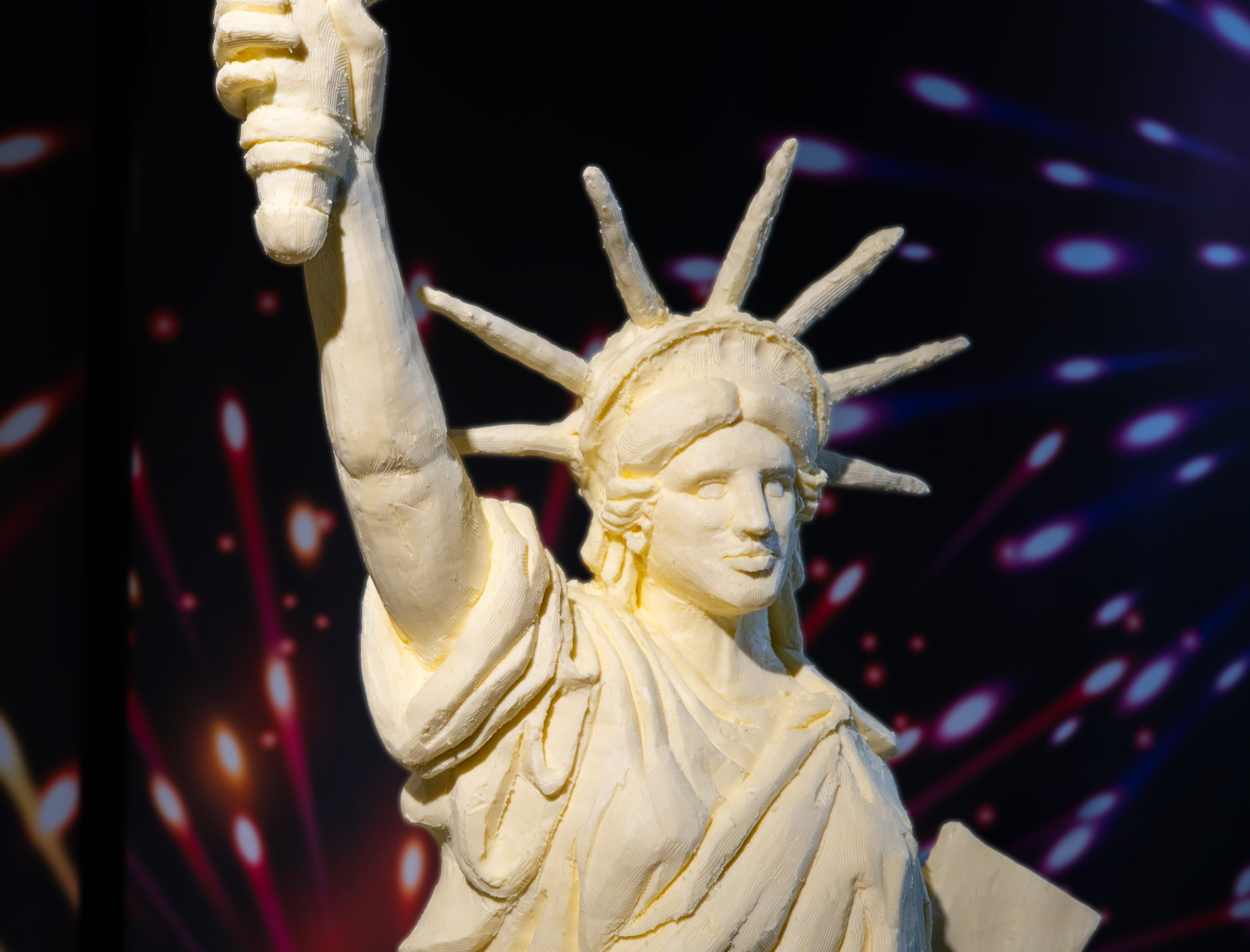 A close-up of the face of the Statue of Liberty carved out of butter amid a backdrop of fireworks.