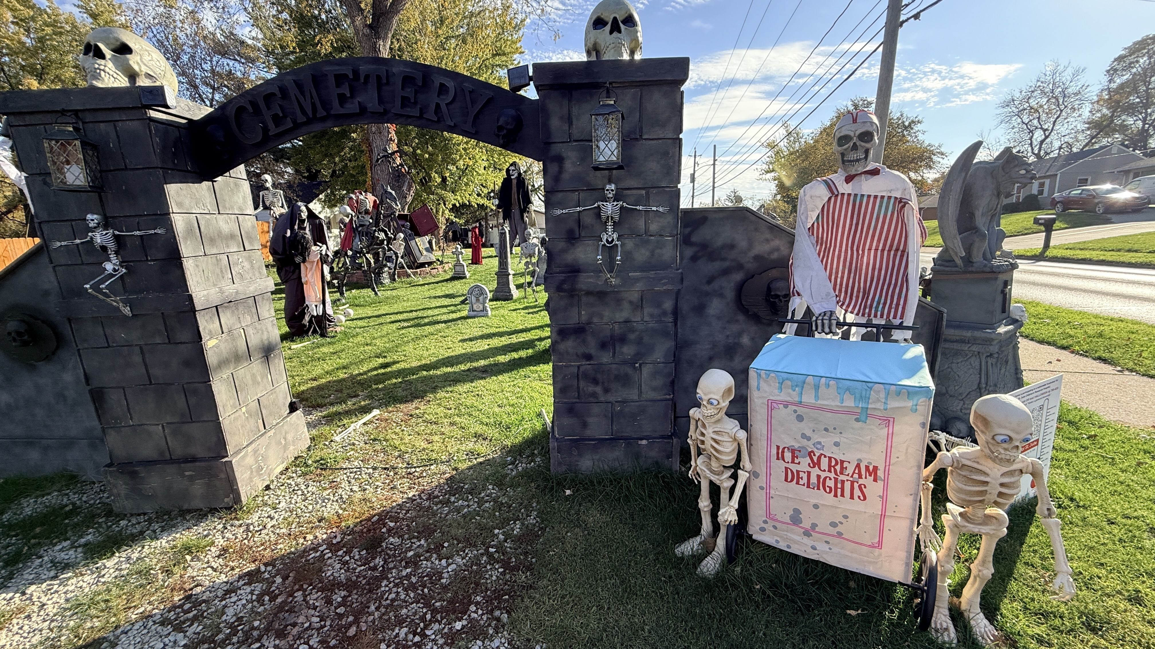 Wide Halloween display: gray stone cemetery gate with skulls, several skeletons posed around, including a striped-suit figure; a gargoyle statue and a sign reading "Ice Scream Delights" nearby.