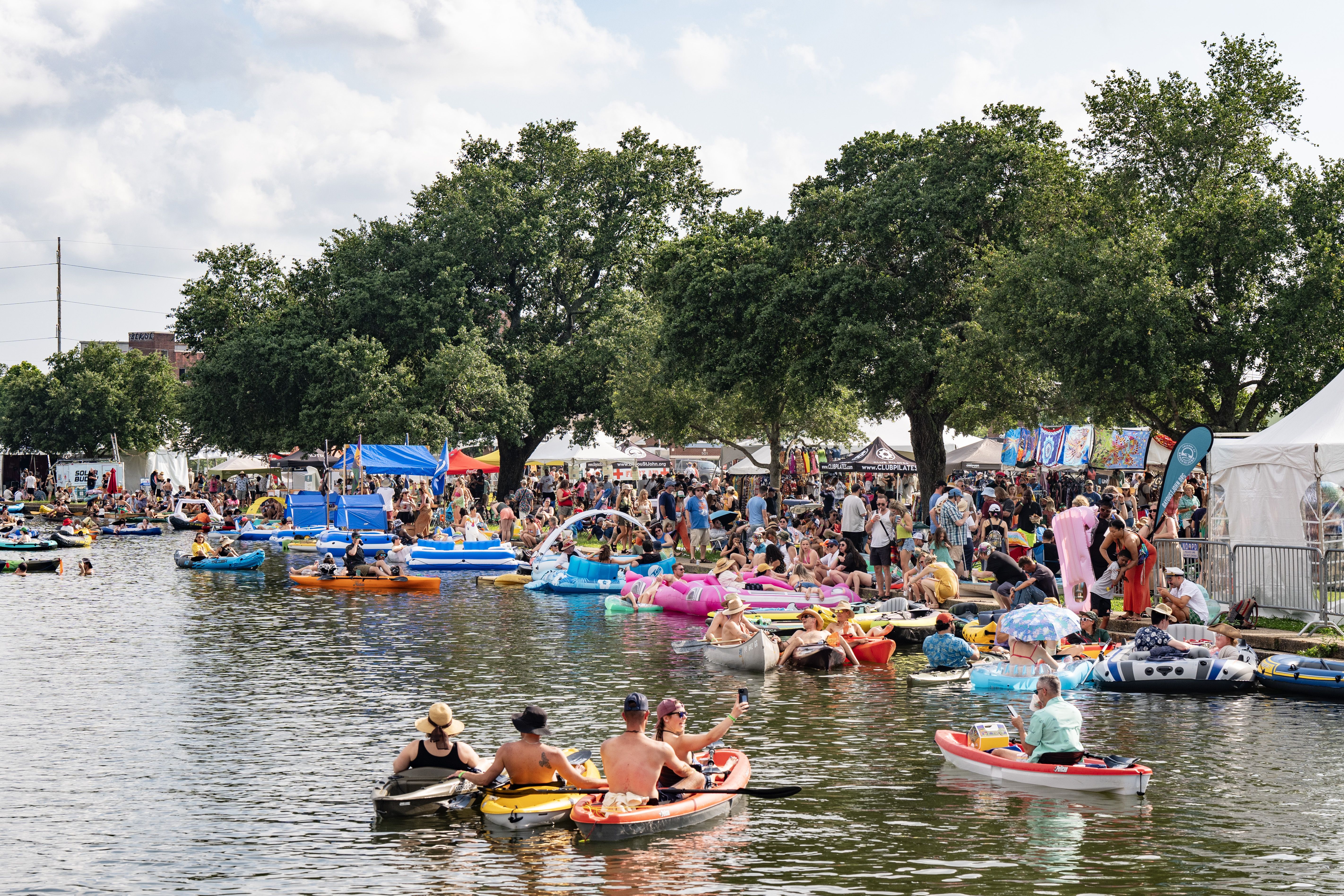 A bayou is filled with boats, floaties and kayaks as a festival takes place at the water's edge.