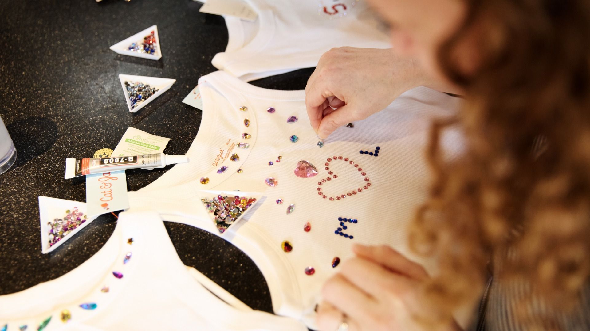 Overhead view of a person decorating a white T-shirt with colorful rhinestones, forming a heart design; small triangular dishes hold gems and a glue tube on a speckled black countertop.