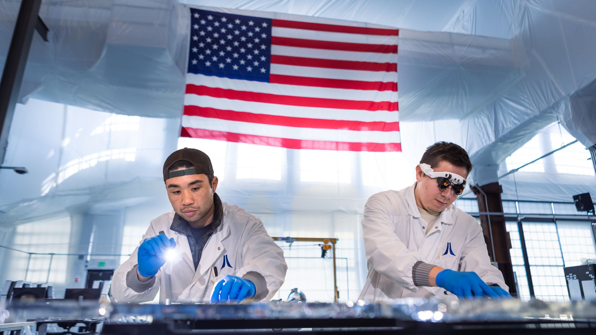 Two men in white lab coats and blue gloves work at a bench beneath a large hanging American flag in a bright, blue-toned laboratory.