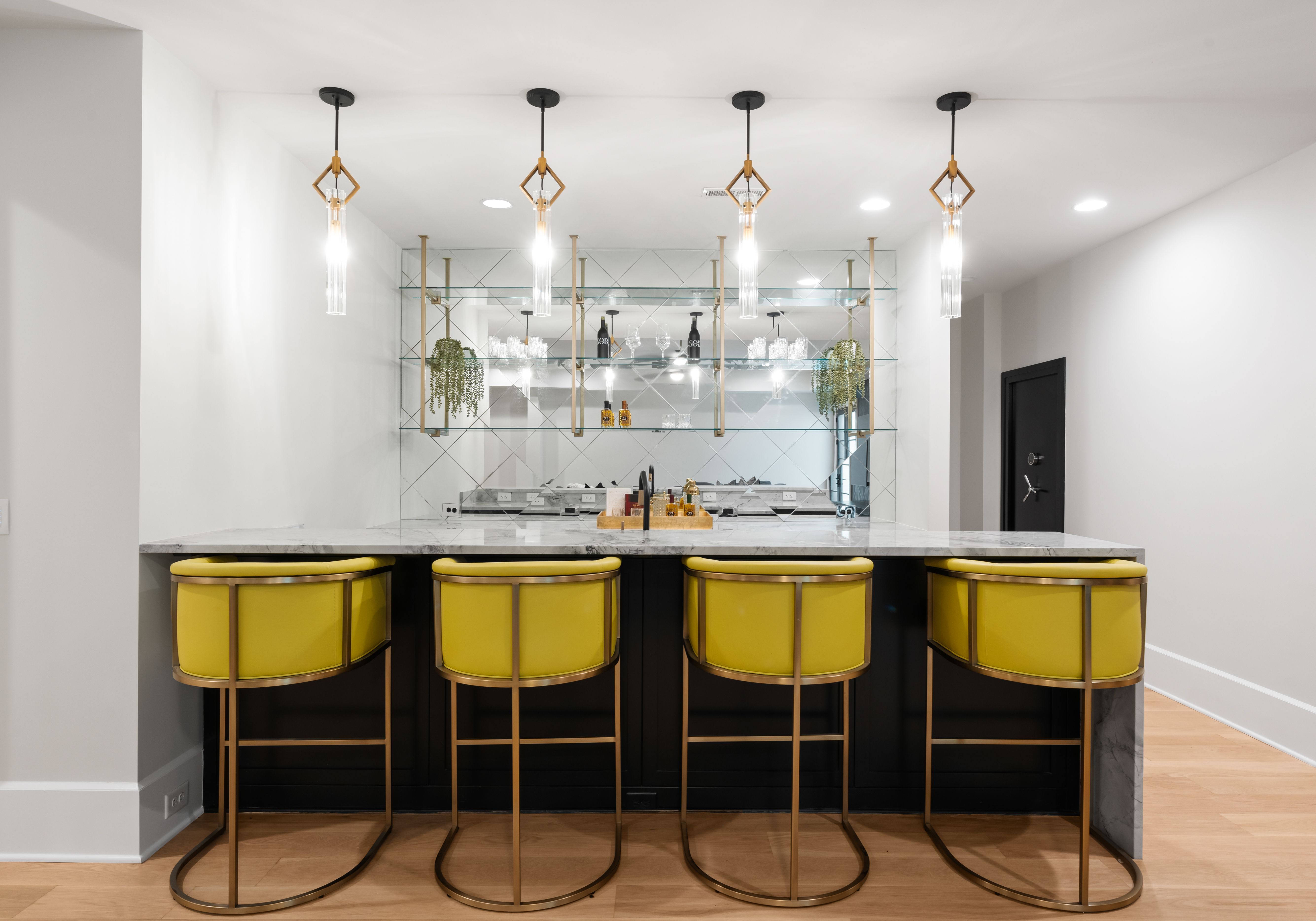 Modern home bar with four yellow cushioned stools, marble countertop, glass shelves with decorative items, hanging pendant lights, and mirrored backsplash in bright room.