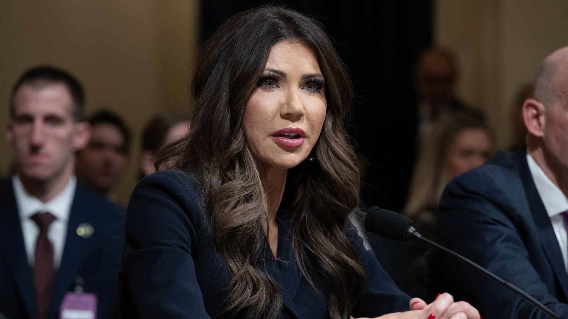A fair-skinned, heavily made up Homeland Security Secretary Kristi Noem, who has dyed dark brown hair stares with her green eyes as she speaks into a black mic as she clasps her hands with nails painted scarlet and wears a navy jacket during congressional testimony.