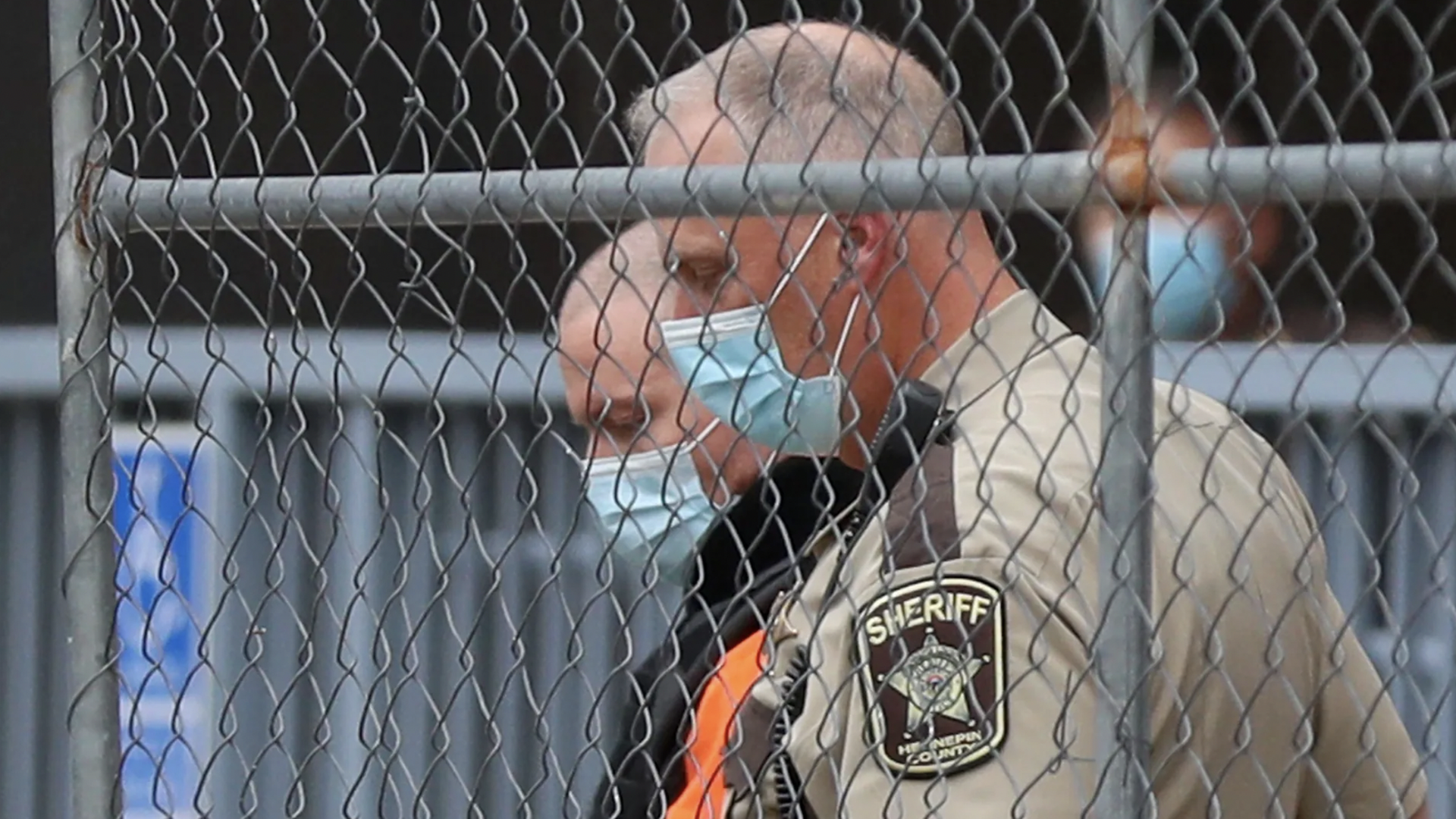 Derek chauvin in a mask being escorted by a sheriff's officer behind a fence