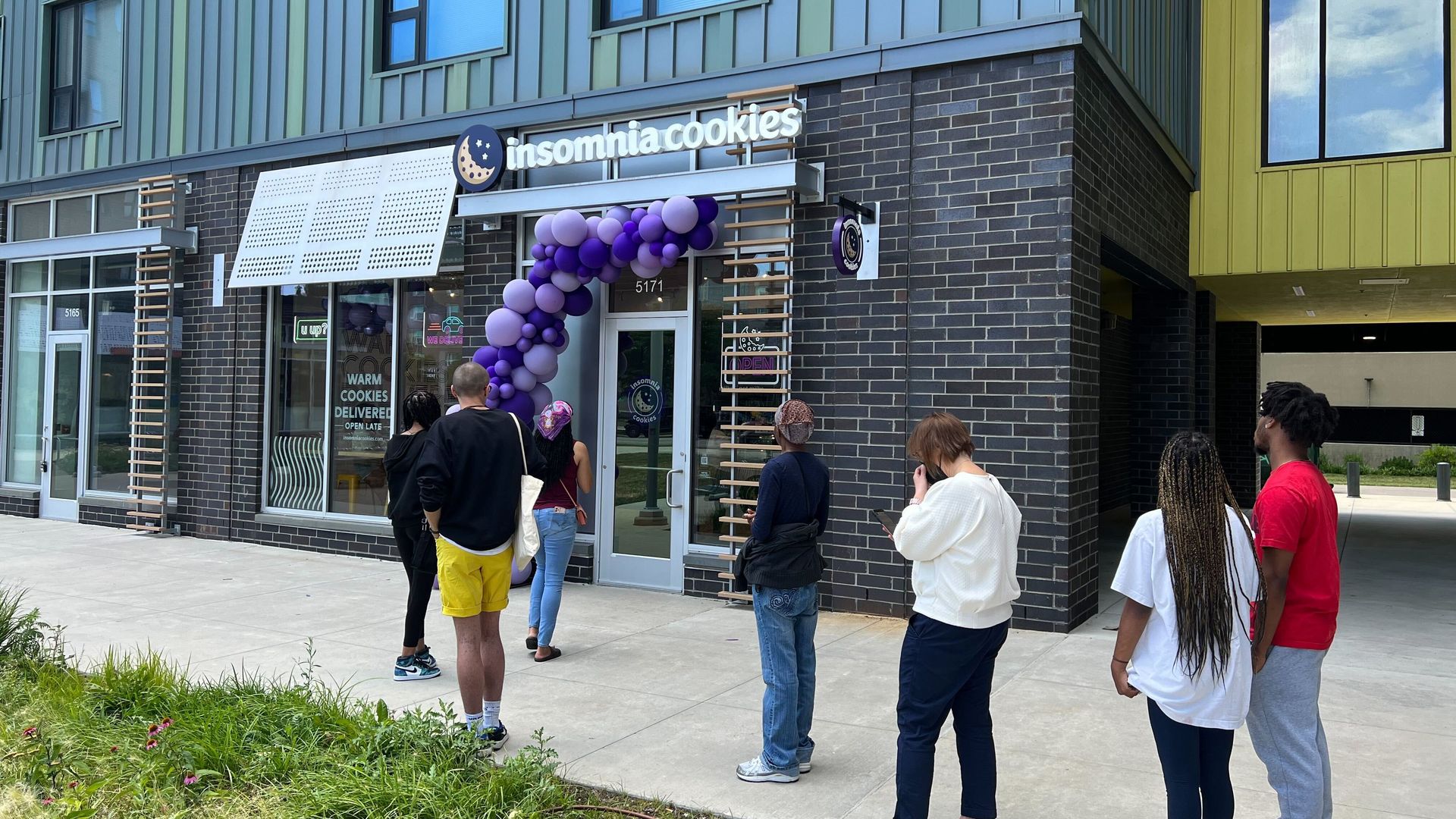 People wait in line outside the storefront for Insomnia Cookies, which is decorated with balloons.